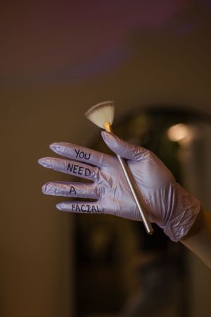 Gloved hand with "You Need A Facial" written on fingers holding a fan brush.