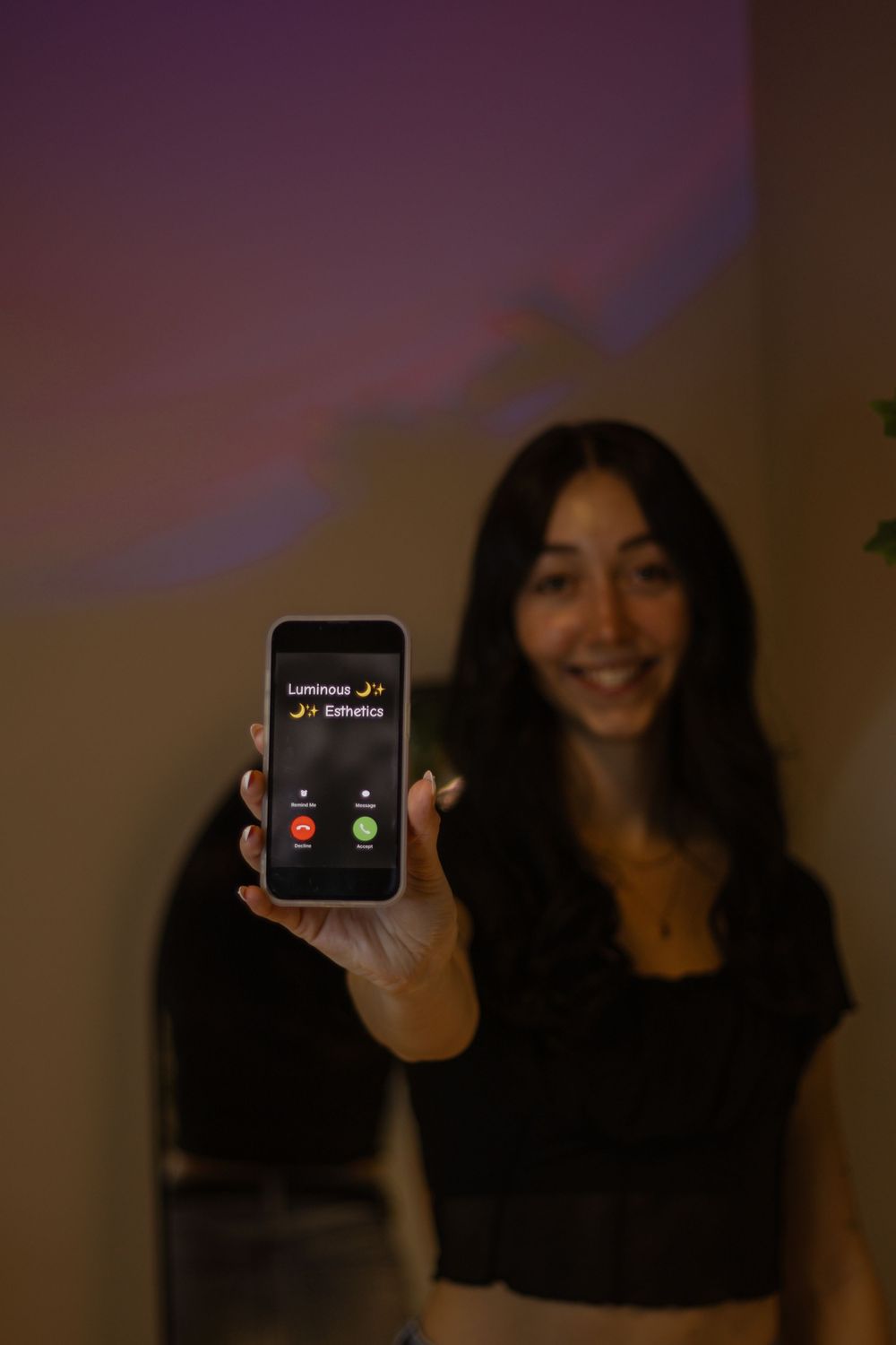 Smiling woman holding phone showing incoming call from Luminous Esthetics in dimly lit room.