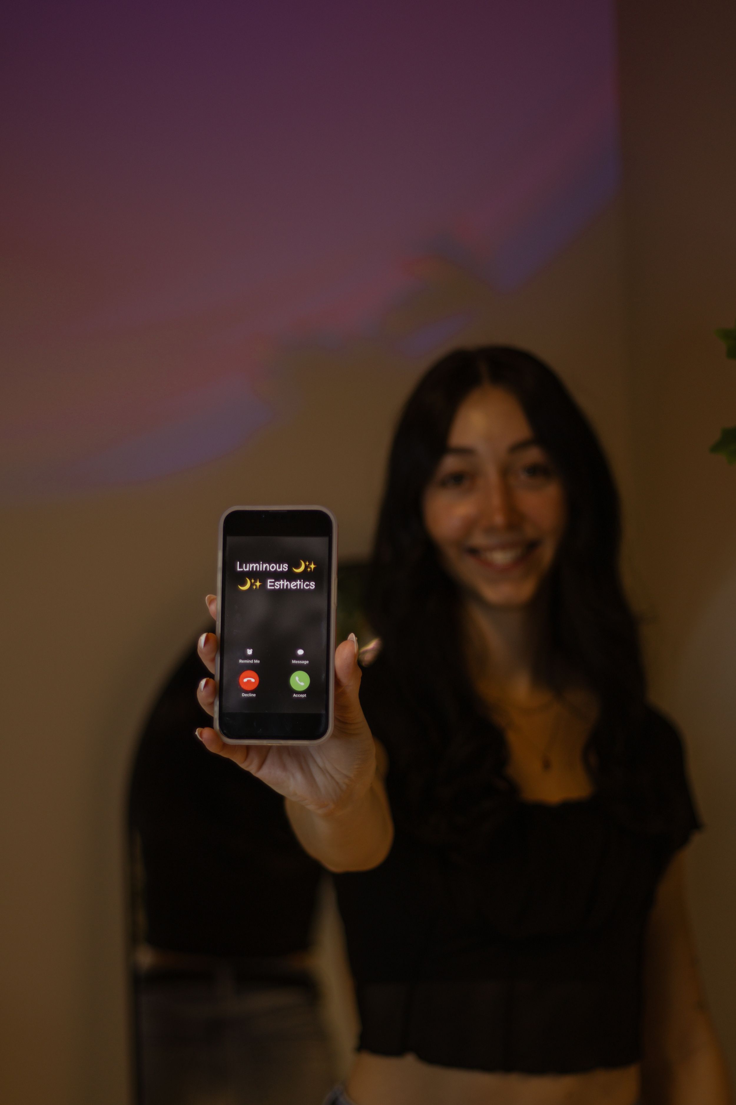 Smiling woman holding phone showing incoming call from Luminous Esthetics in dimly lit room.