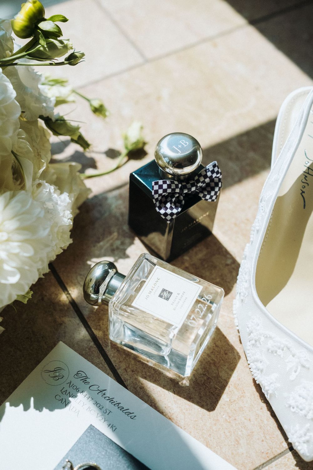 Bridal flat lay with Jo Malone perfume bottles, white floral bouquet, lace heels, and wedding stationery in sunlight.