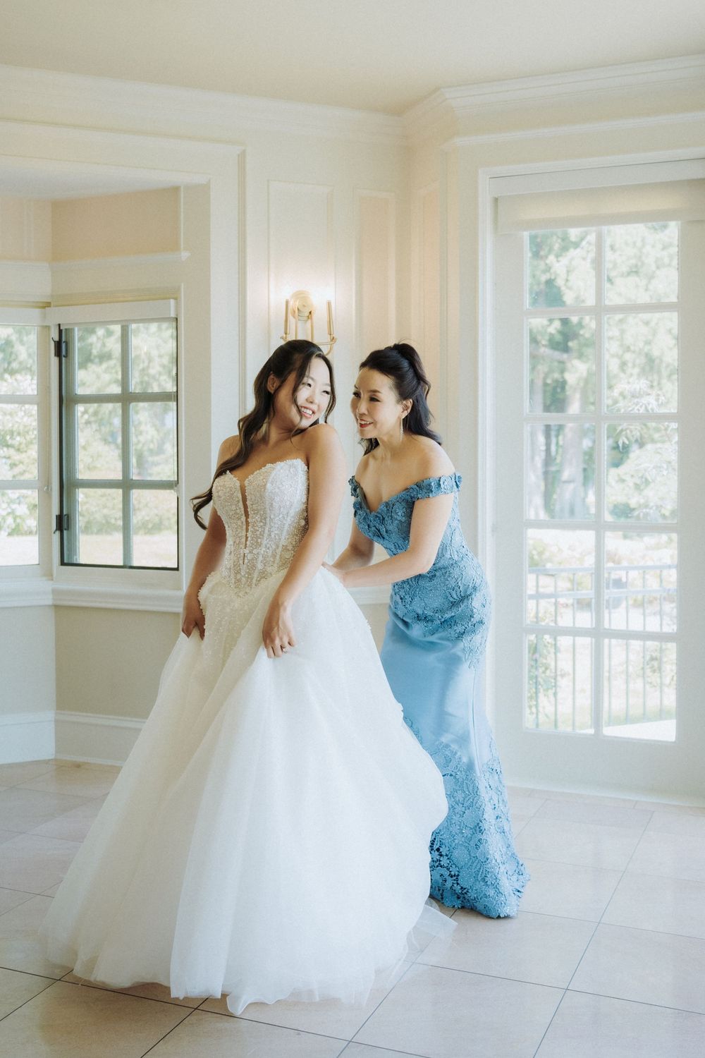 Bridesmaid in blue lace gown helps smiling bride adjust her strapless ballgown in a bright elegant dressing room.