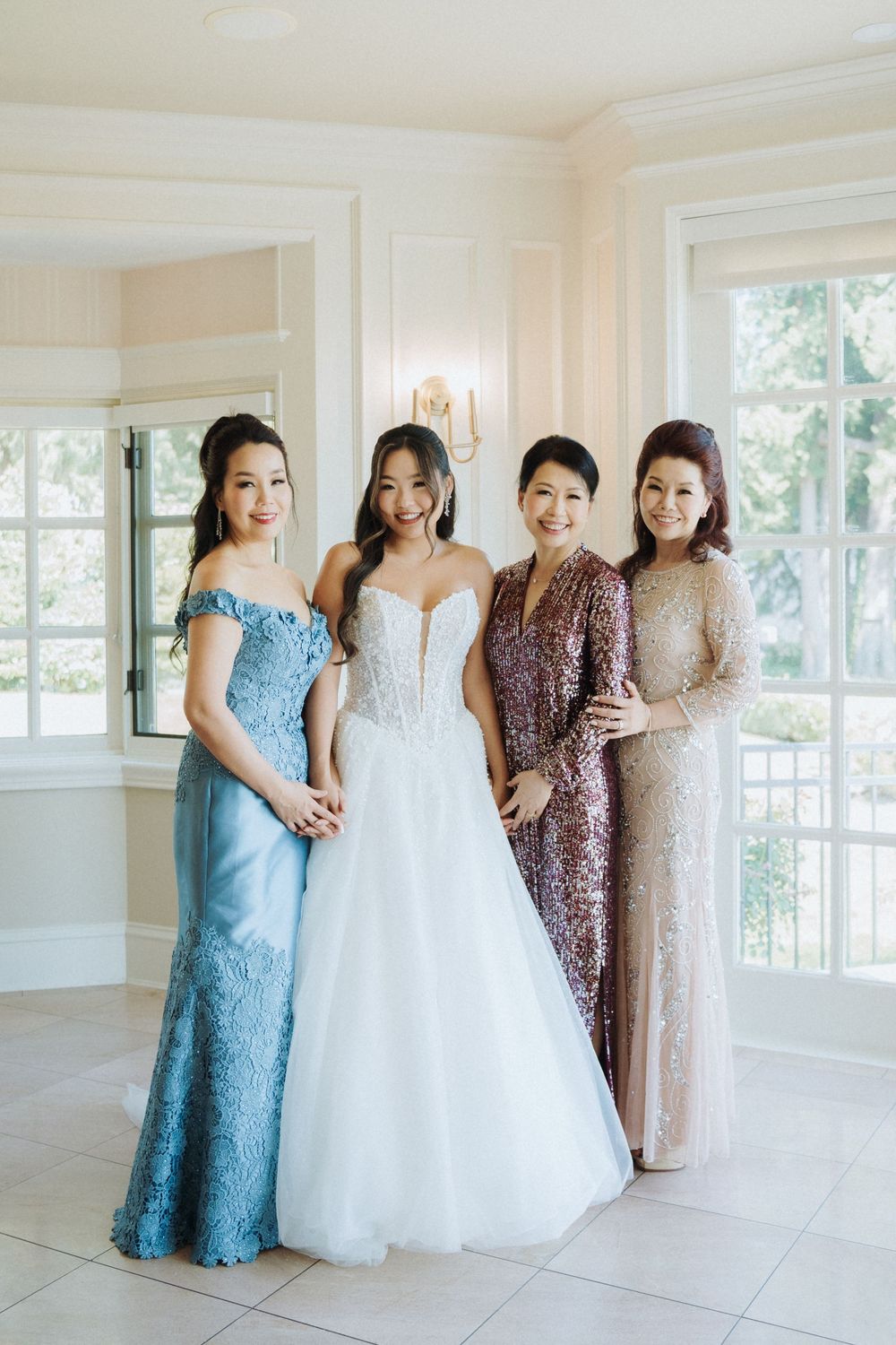 Bride in white ballgown poses with three elegantly dressed women in a bright, airy room before the wedding.