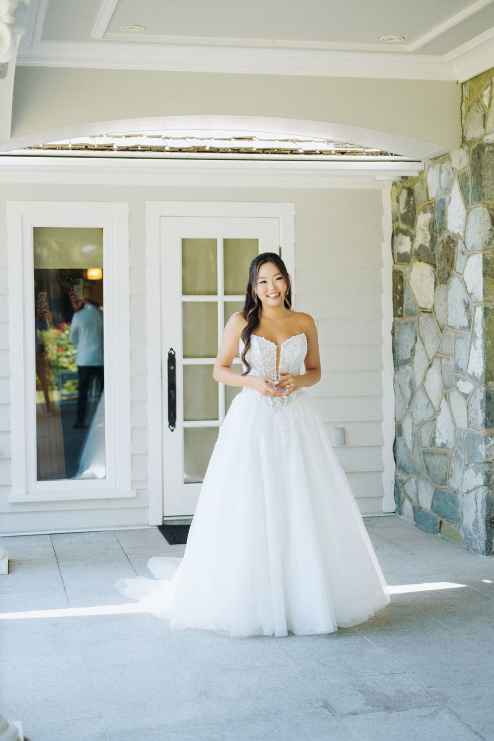Bride in strapless lace ballgown wedding dress stands smiling outside white venue with stone wall.