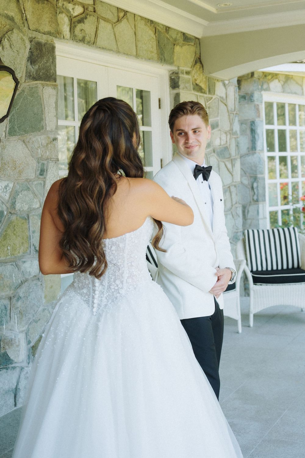 Groom in white tuxedo jacket smiles at bride in strapless lace wedding gown during outdoor first look moment.