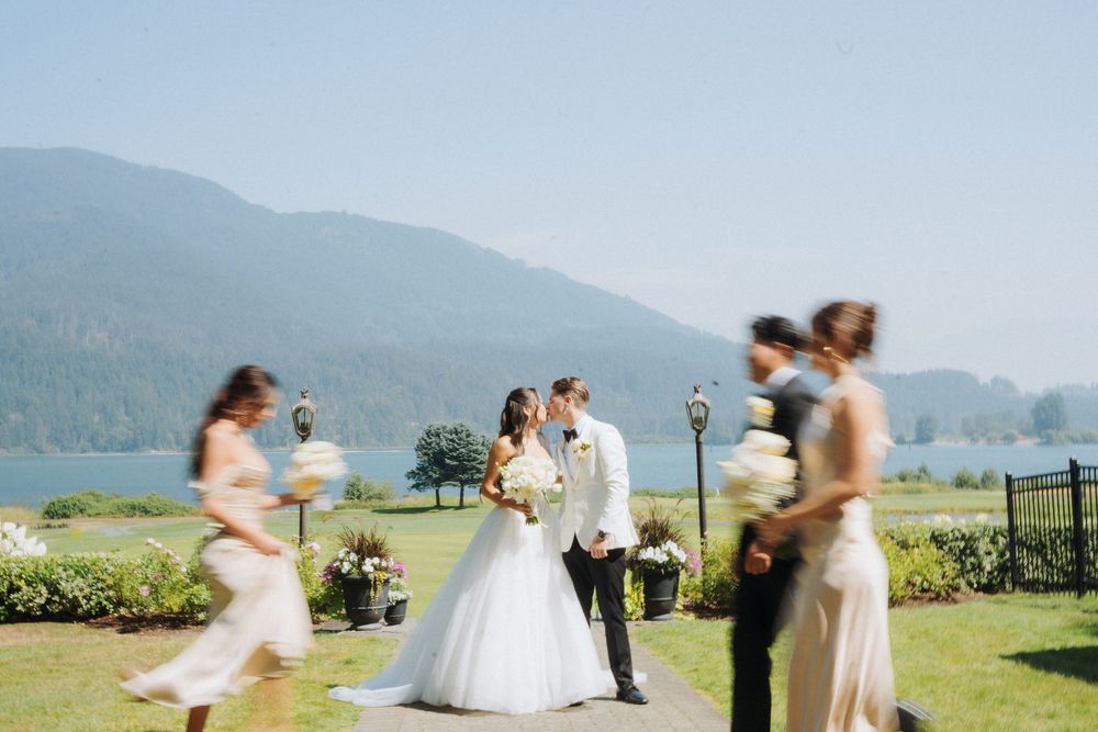 Bride and groom kissing outdoors with mountain lake backdrop as bridesmaids walk past in elegant gowns.