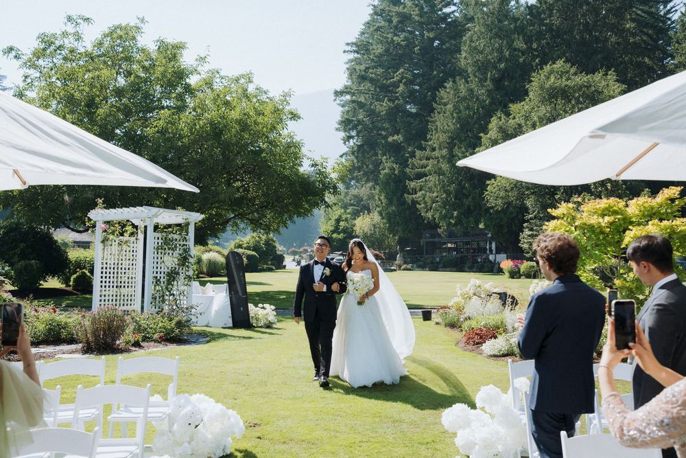 Bride in white gown walks down outdoor aisle with escort during sunny garden wedding ceremony.
