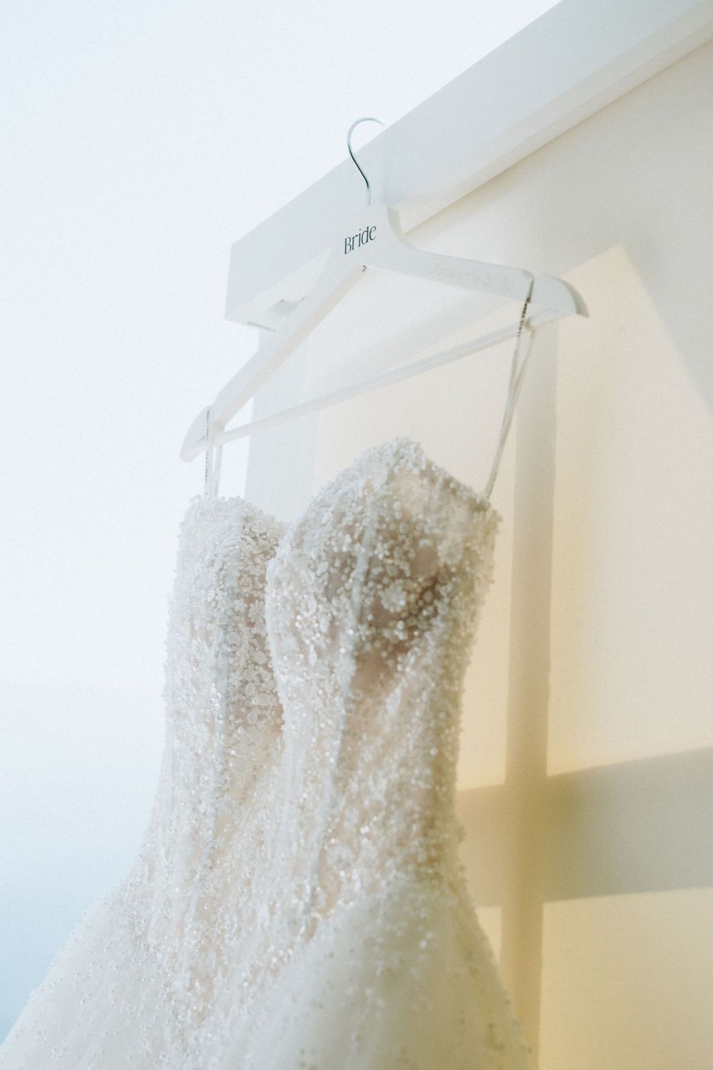 Beaded strapless wedding gown hanging on white bride hanger against soft white background.