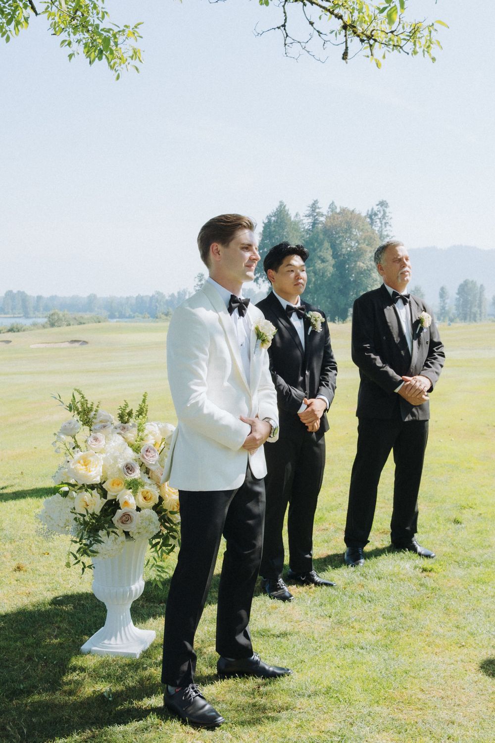 Groom in white tuxedo stands at outdoor wedding altar with two groomsmen on a sunny golf course ceremony.