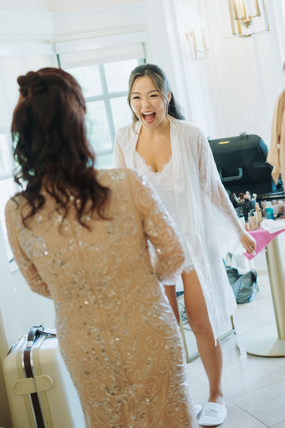 Joyful Asian bride in white lace robe laughing with guest in sequin dress during wedding prep.