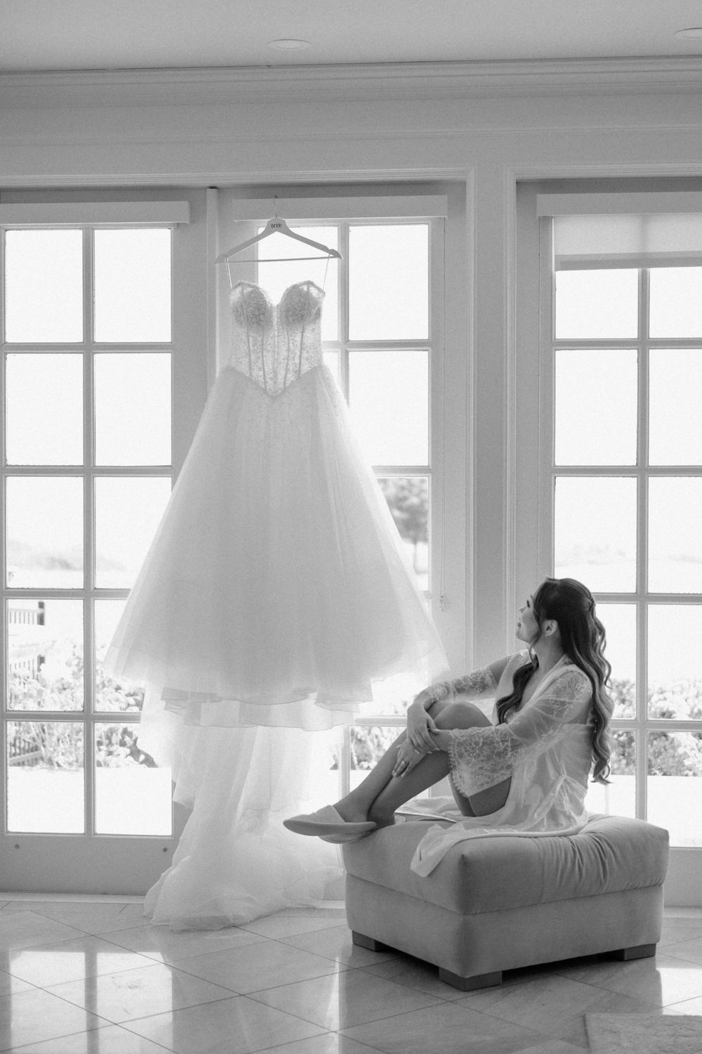 Bride in robe gazing at elegant beaded ballgown wedding dress hanging by sunlit French windows in black and white photo.