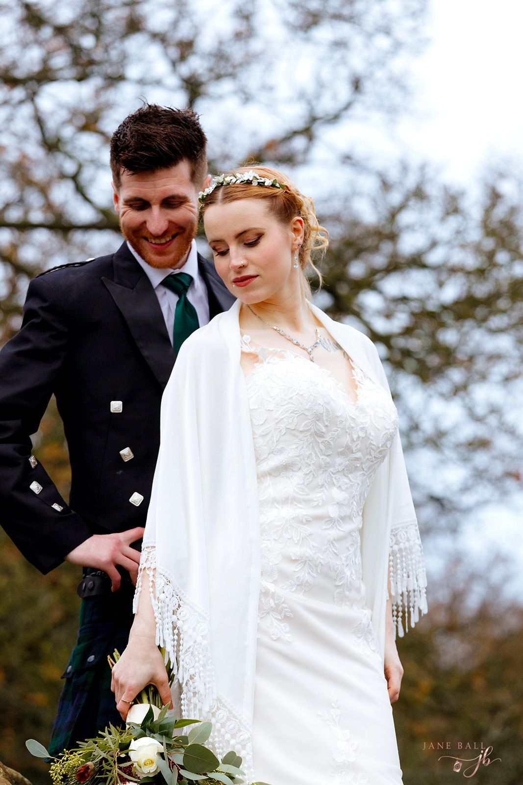 Bride in white lace gown and fringe shawl with groom in Scottish kilt smiling outdoors at winter wedding. The Outbarn