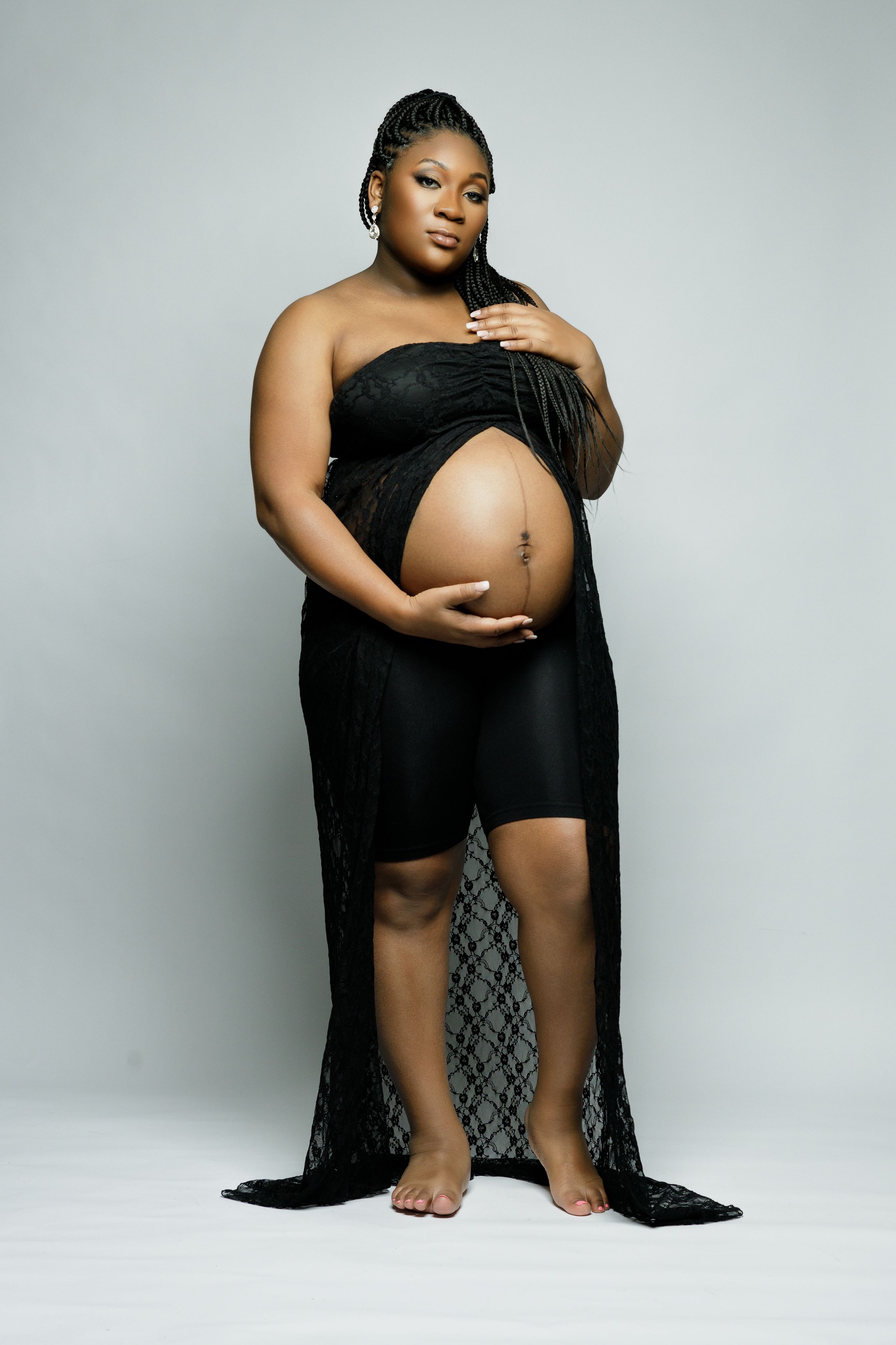 Artistic maternity portrait showing pregnant woman in black outfit with lace overlay against light background.