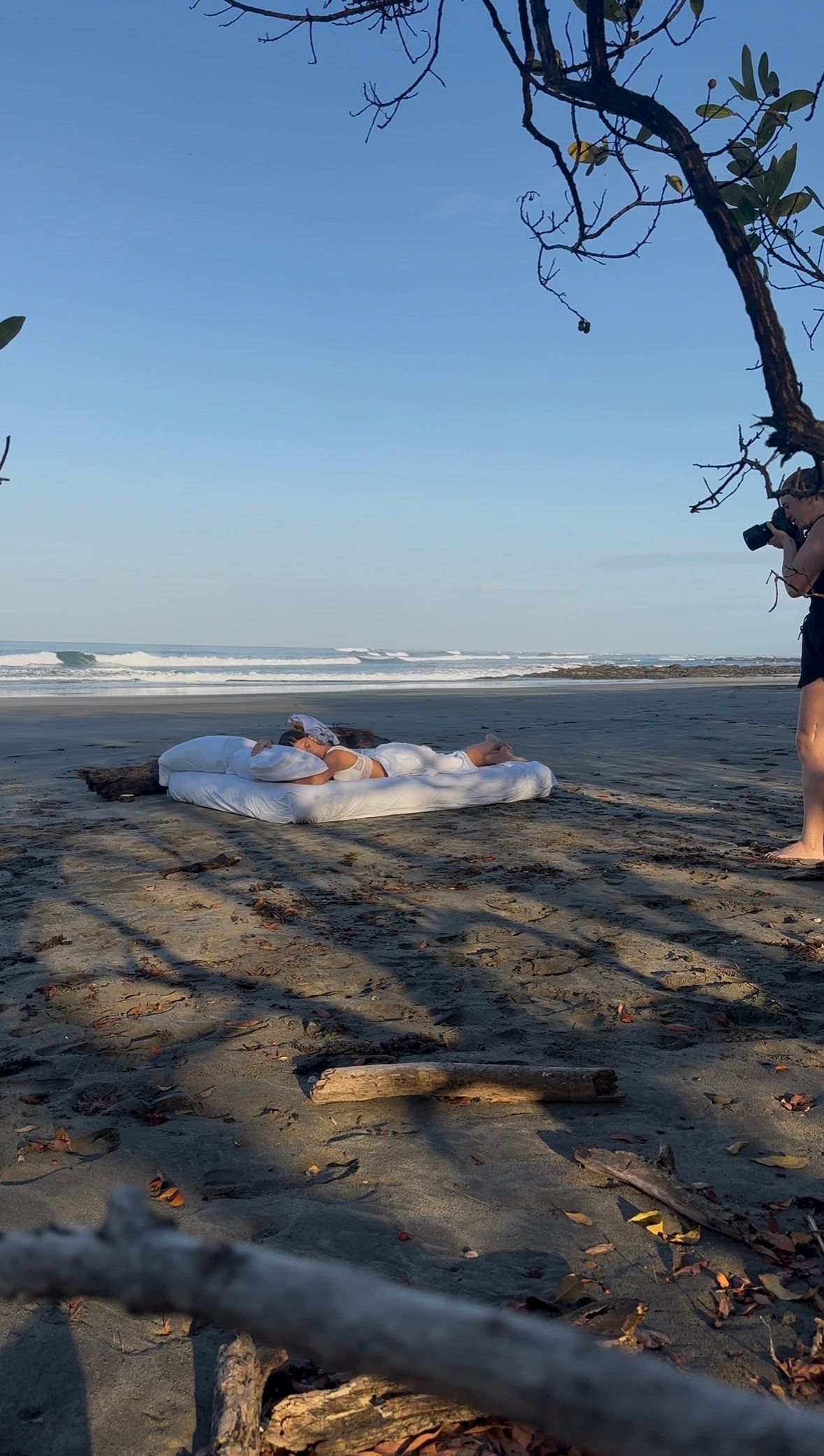 Two people lying on a white mattress on a sandy beach with ocean waves and clear blue sky in the background.