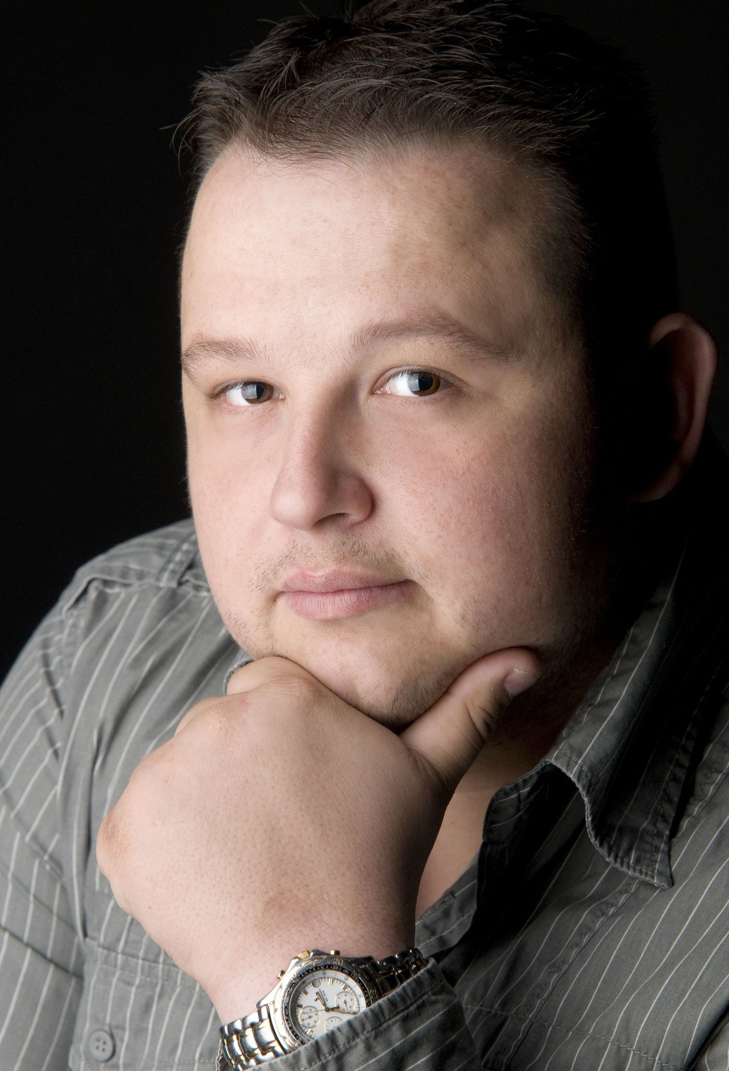 Professional portrait photograph of a person in a striped button-down shirt with a silver watch, posing with hand on chin.