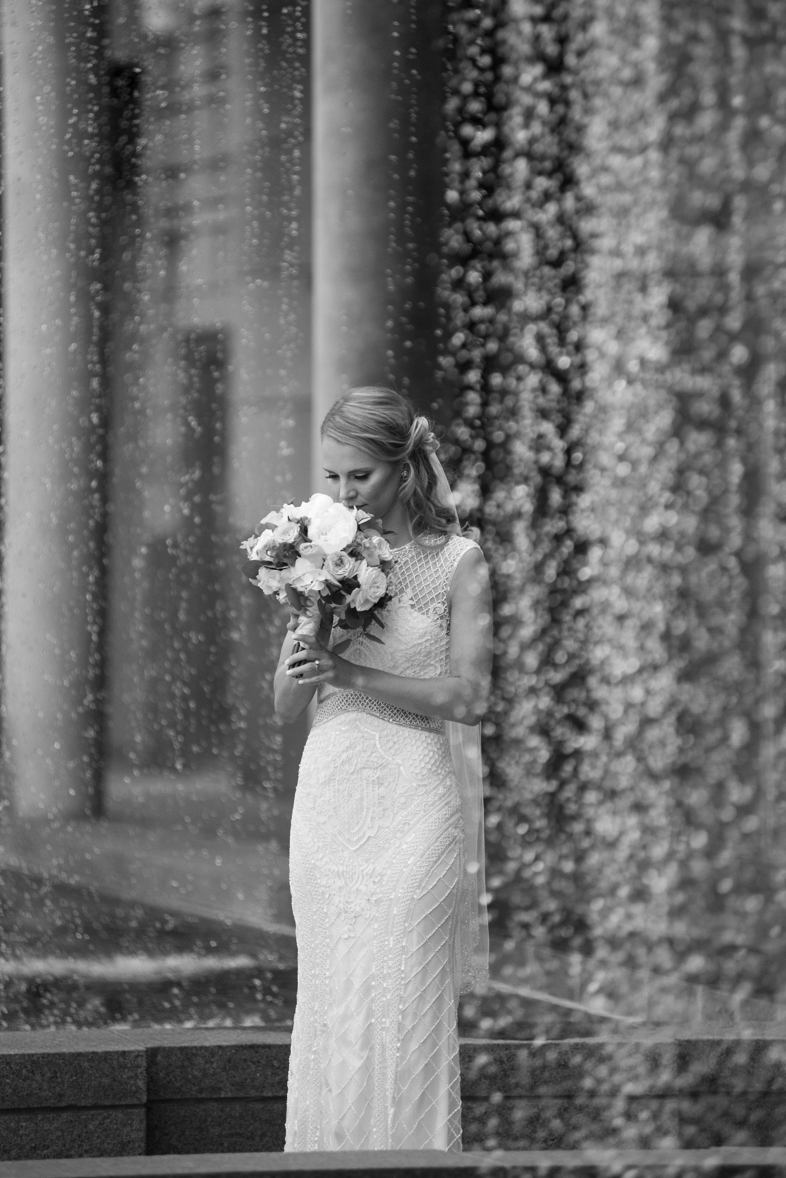A bride in a white lace dress holds a bouquet while standing against a backdrop of cascading floral vines.