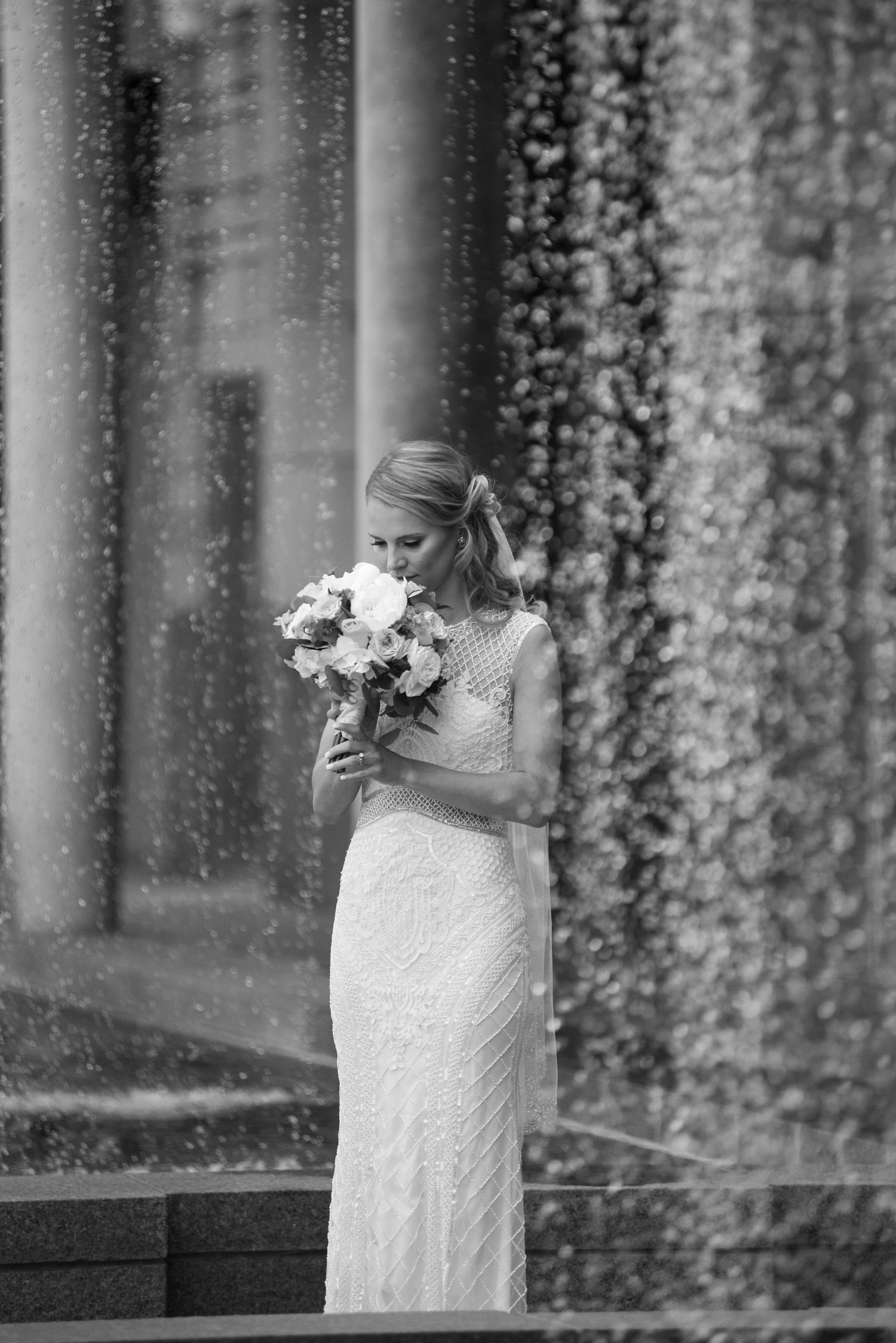 A bride in a white lace dress holds a bouquet while standing against a backdrop of cascading floral vines.