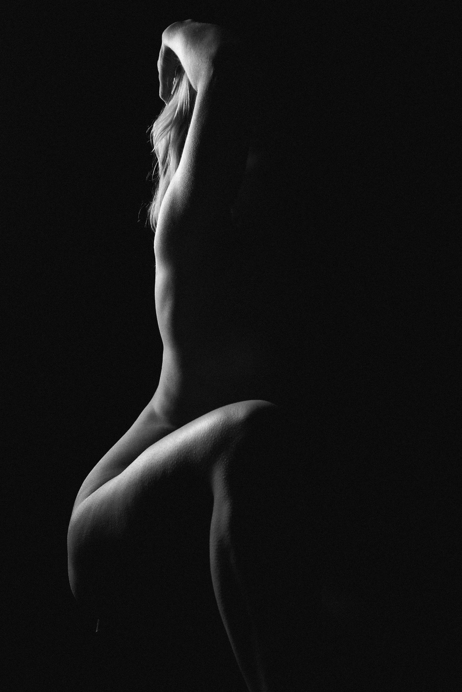 Artistic black and white silhouette of a nude woman seated against a dark background.