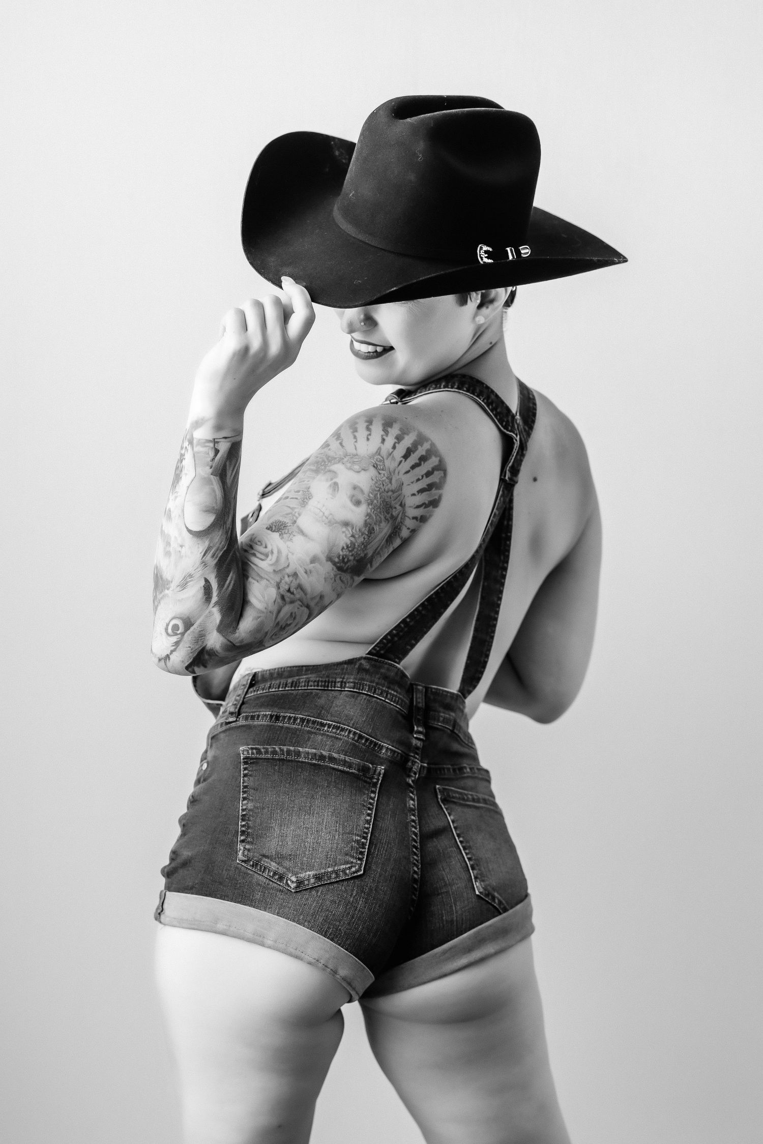Tattooed woman in denim shorts and black cowboy hat posing from behind in black and white photo.