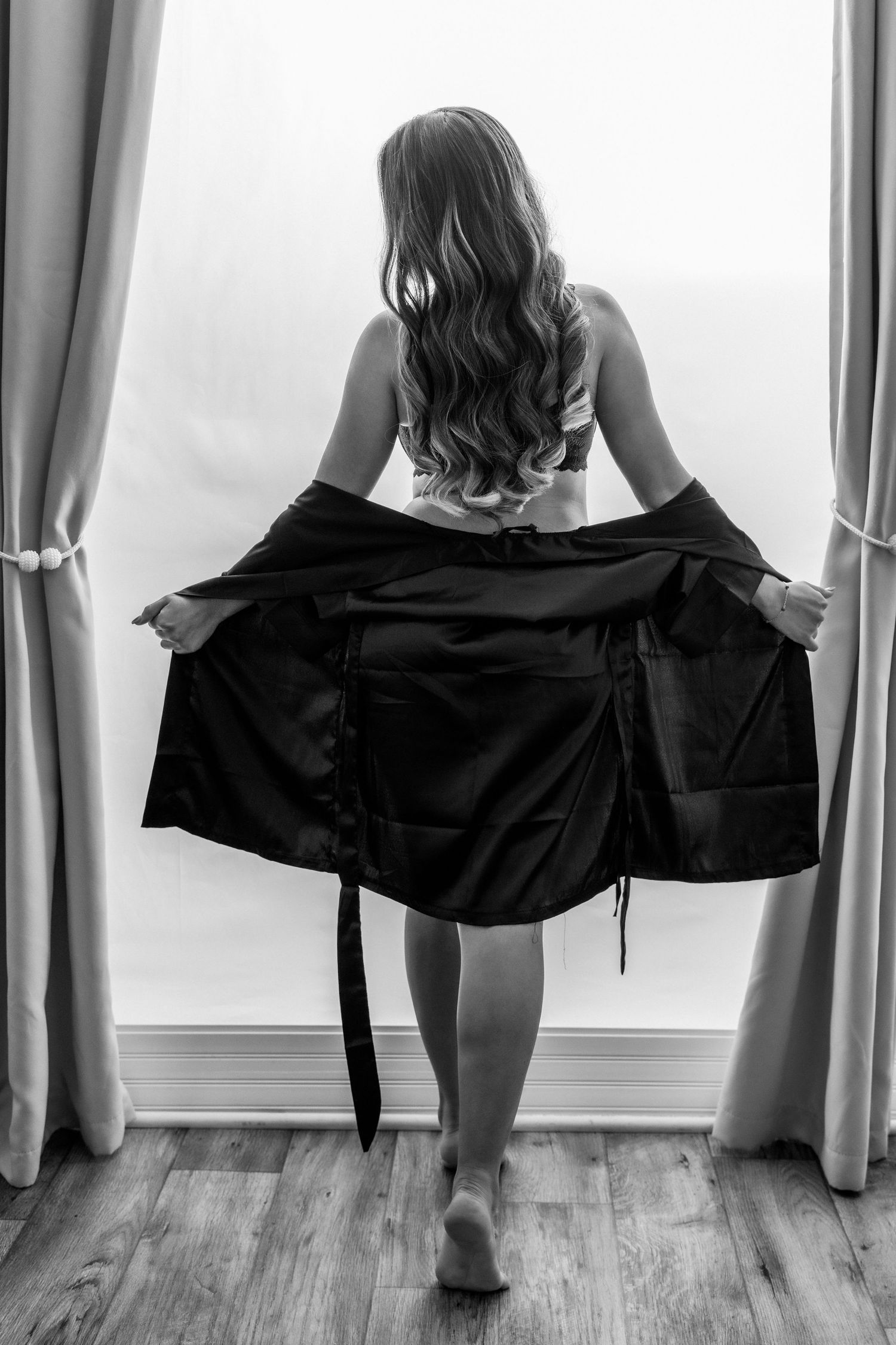 Black and white rear view of woman in white bra holding open robe near curtained window.