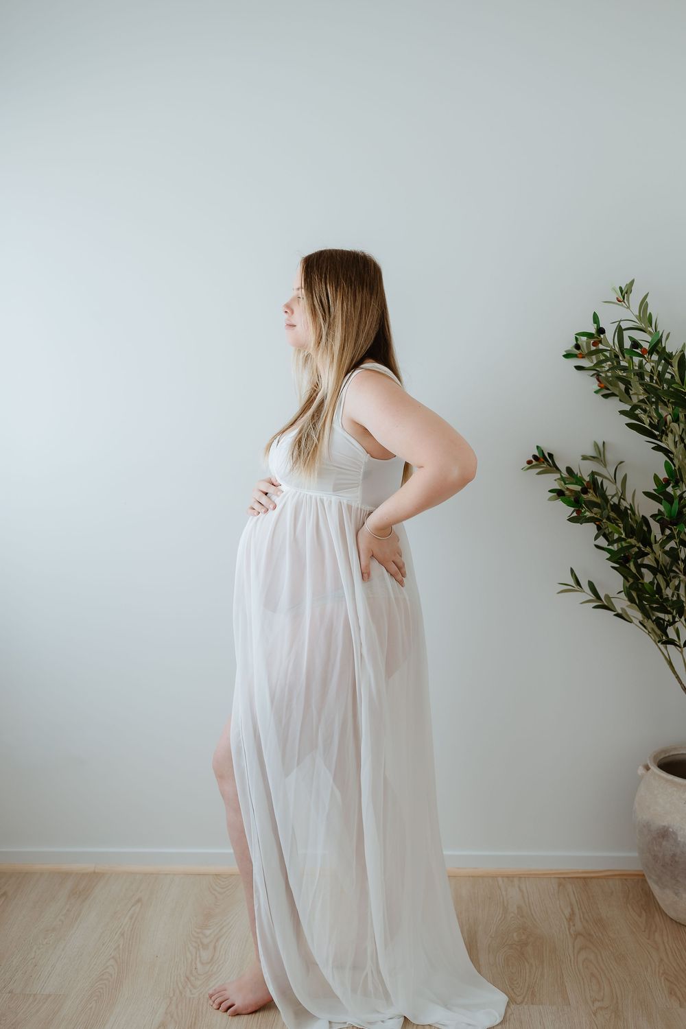 Expectant mother in flowing white maternity gown poses elegantly against light wall with potted plant in minimalist setting.