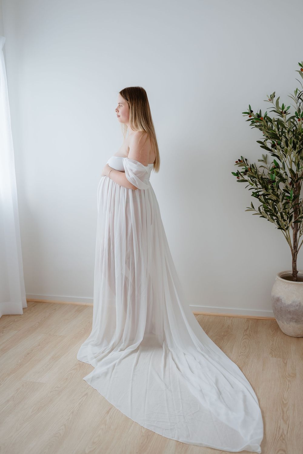 A flowing white maternity gown with off-shoulder design is showcased in multiple angles on a light wooden floor.
