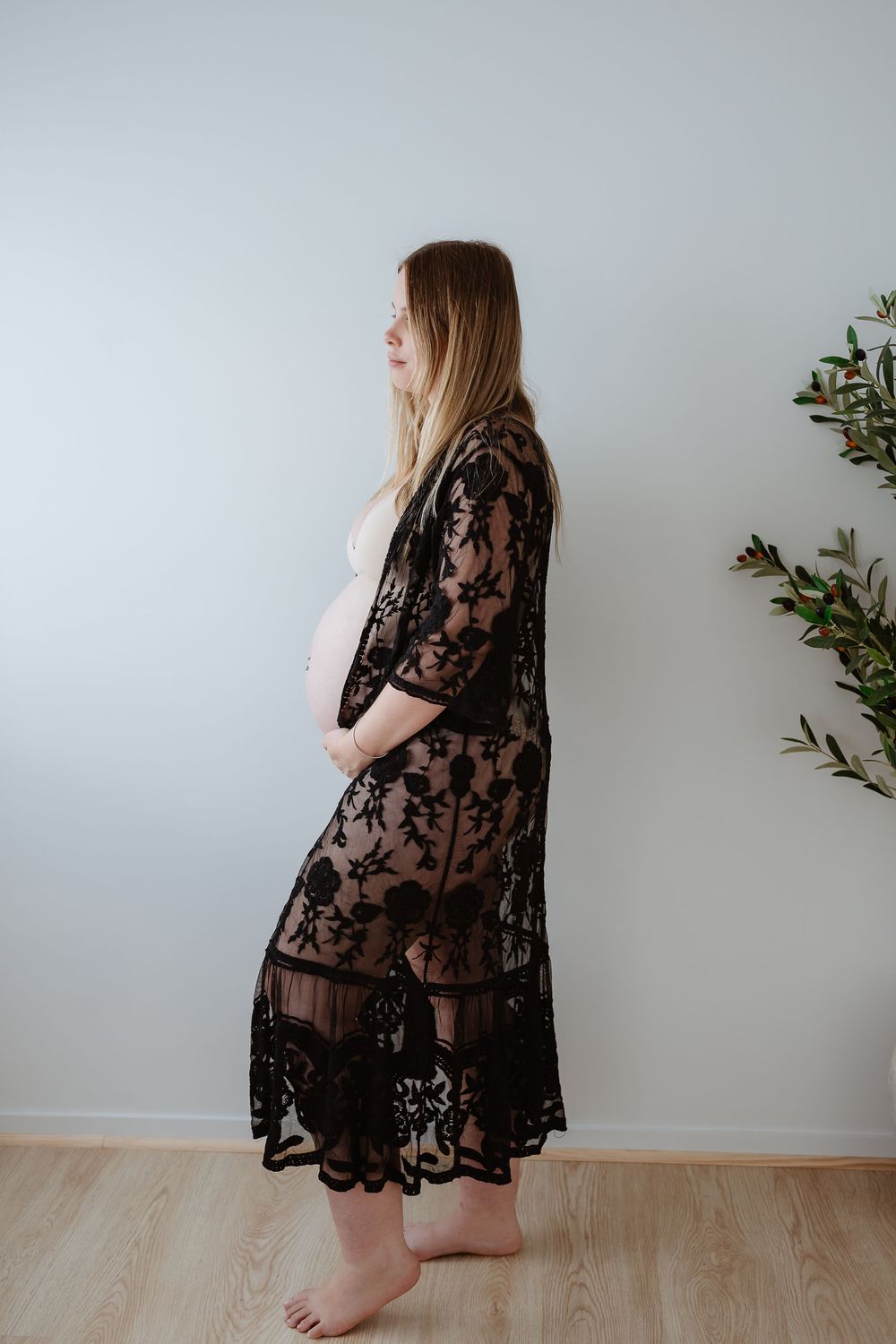 A person wearing a sheer black lace dress poses against a white wall with minimal greenery decoration.