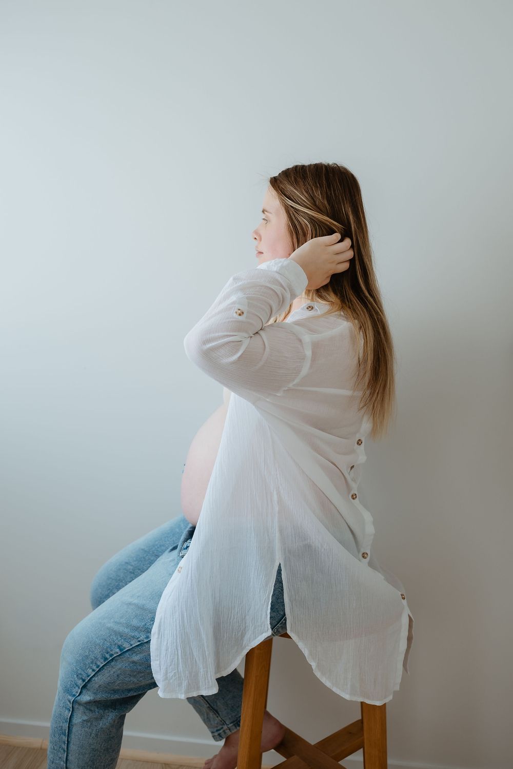 Side view of an expectant mother wearing a flowing white shirt and jeans while sitting on a wooden stool against a light wall.