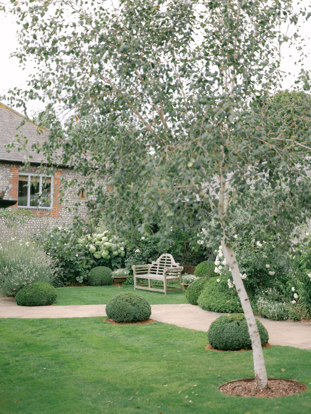 Elegant English garden with silver birch tree, manicured box hedges, white bench, and hydrangeas beside a stone building.
