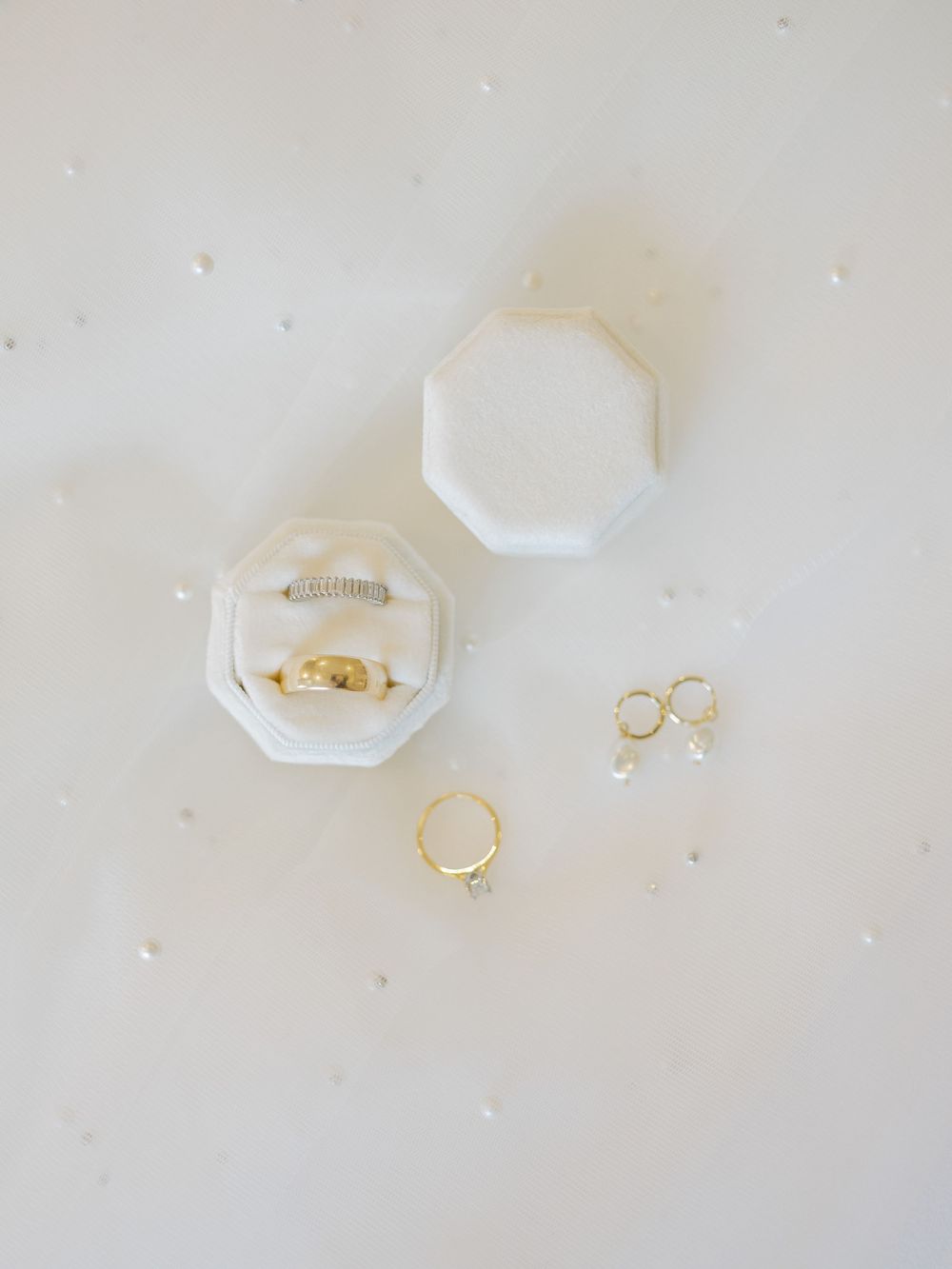Gold and diamond wedding rings in white octagonal velvet ring box with pearl drop earrings on white surface.