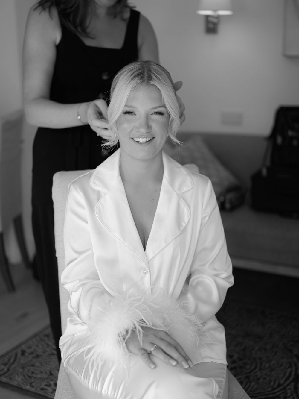 Smiling bride in white silk robe getting hair styled in black and white wedding preparation photo.