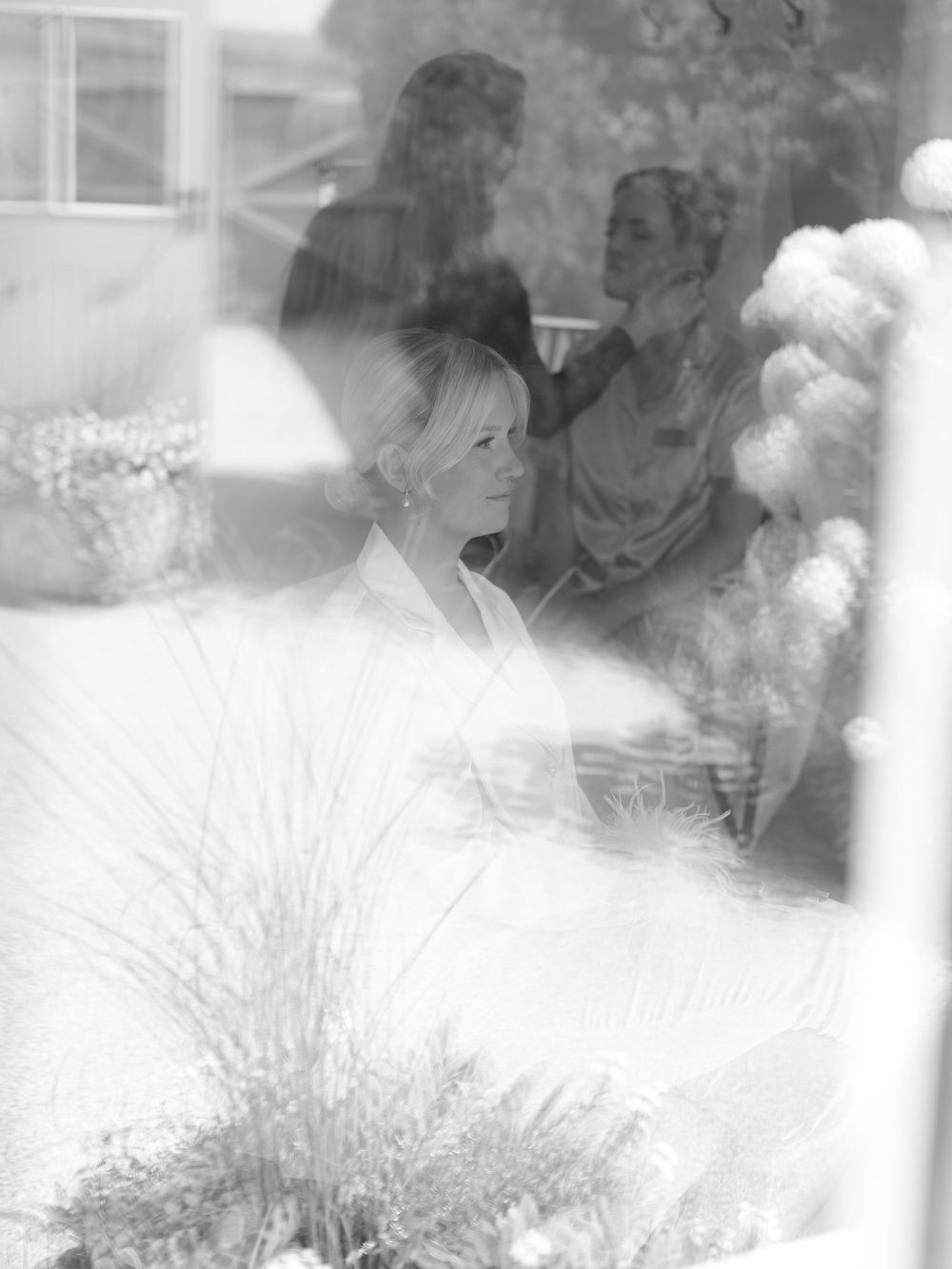 Black and white photo of a blonde woman in white, seen through a window with reflections of people and flowers.