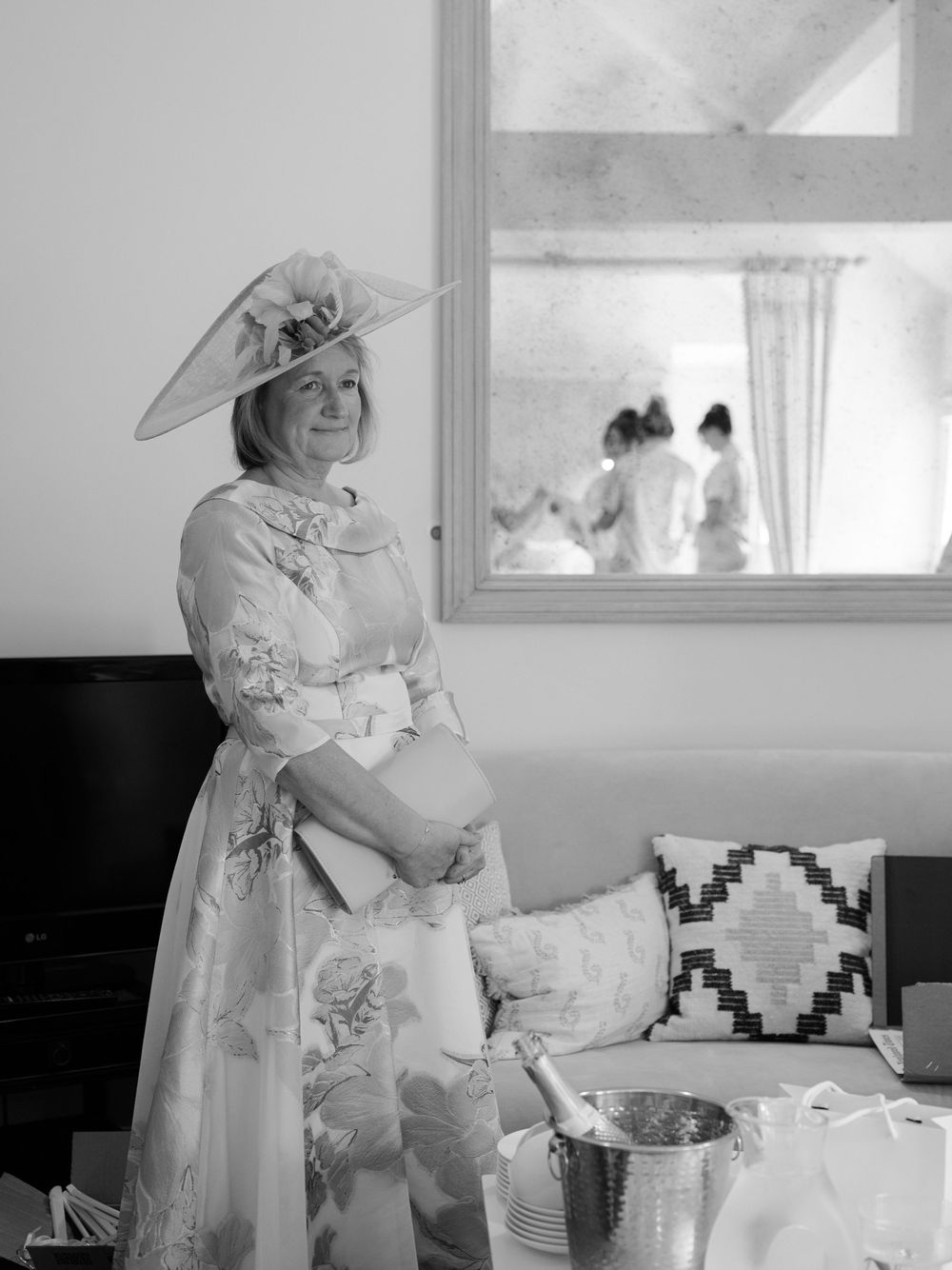 Elegant woman in floral dress and wide-brim hat poses at wedding venue in black and white photo.