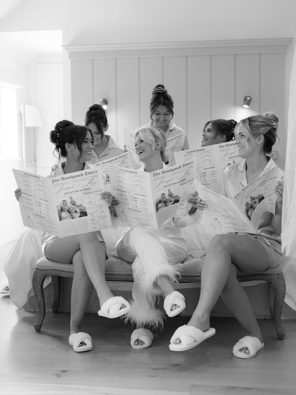 Bride and bridesmaids in robes reading wedding newspapers together on a bench in black and white photo.