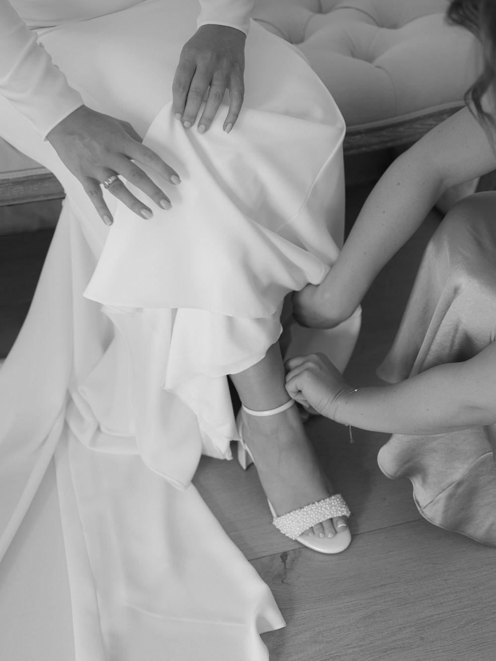 Black and white photo of bridesmaid fastening white heeled sandal on bride in wedding dress.