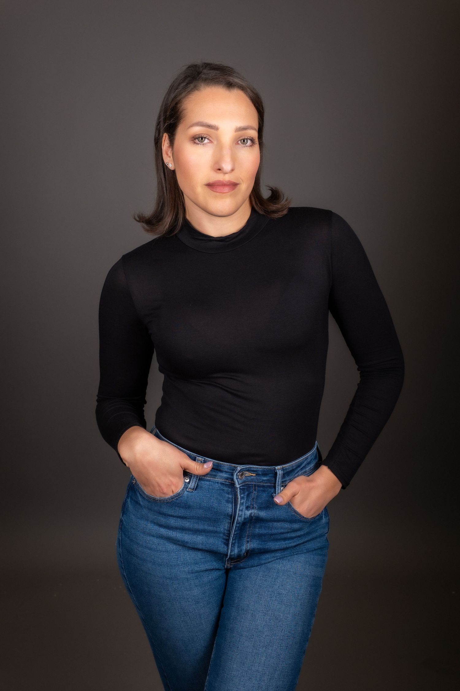 A figure in a black turtleneck and blue jeans poses confidently against a dark studio backdrop.