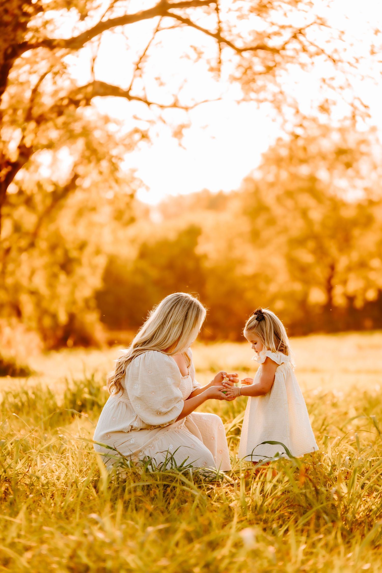 Mother and young daughter in white dresses sharing a tender moment in a golden sunlit field.