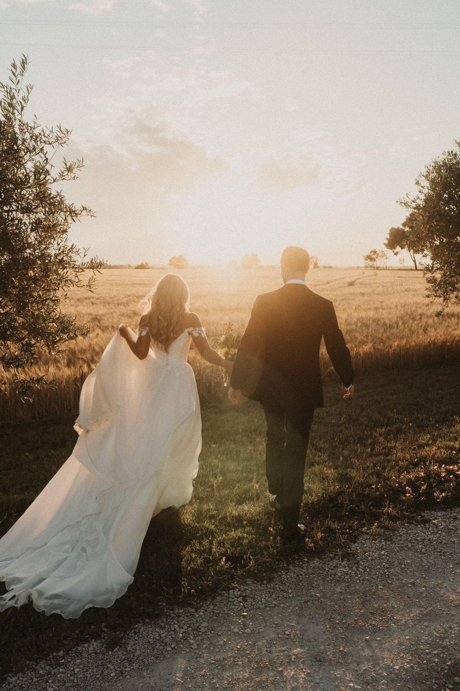 A couple in wedding attire walks along a gravel path at sunset, their silhouettes backlit by golden sunlight.