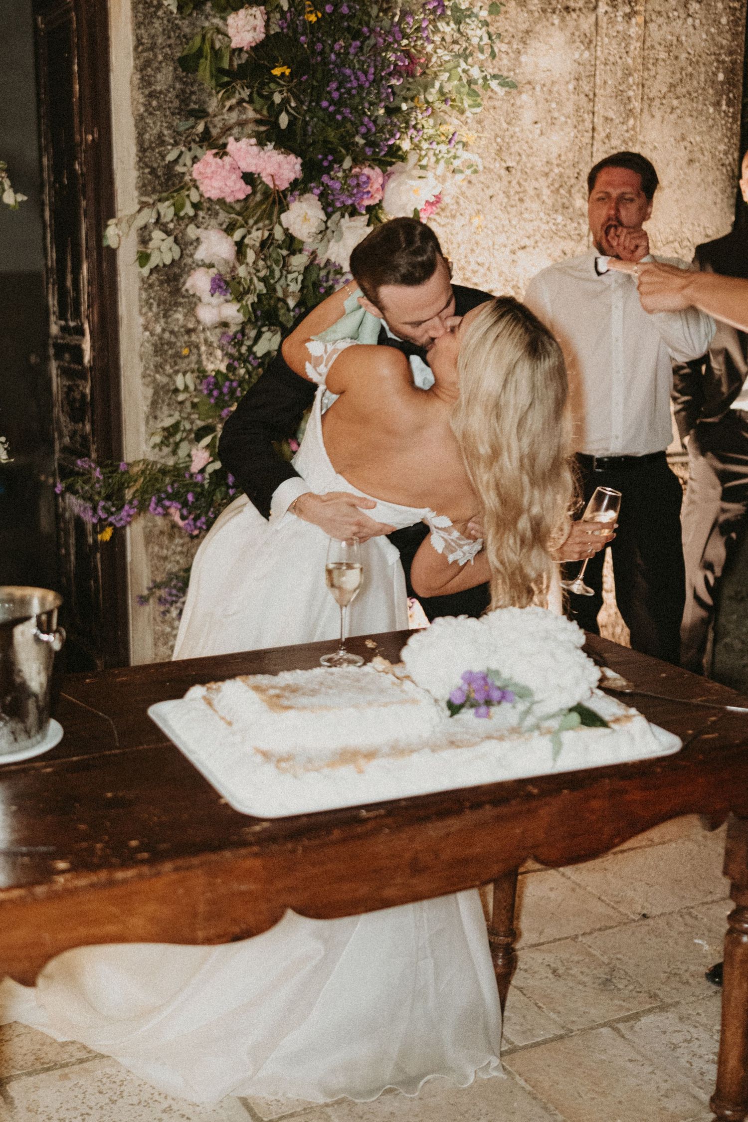 Newlyweds share romantic kiss while cutting their wedding cake at rustic reception with floral decorations.