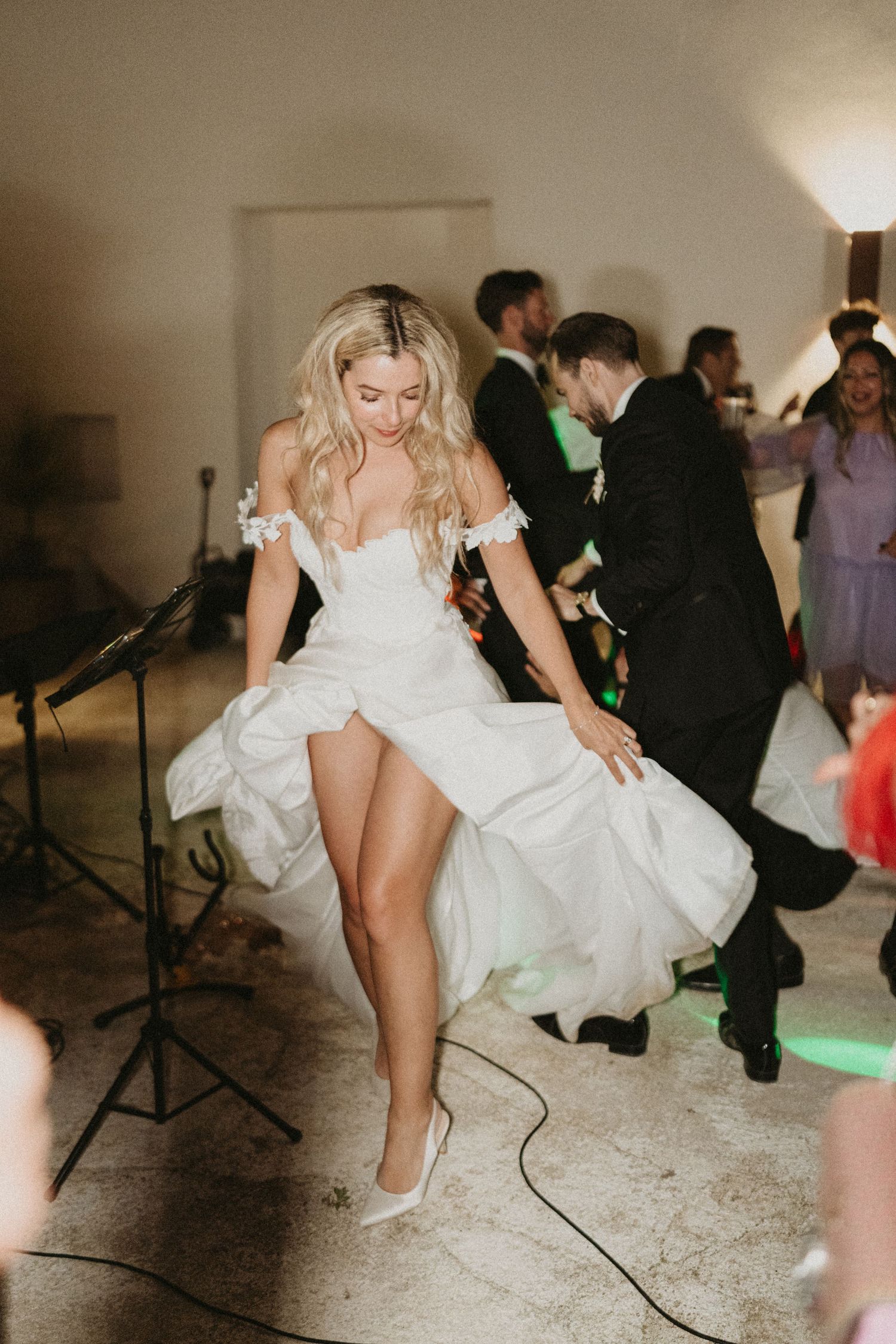A bride in a white wedding dress dances at her reception, lifting her dress while wearing white heels.