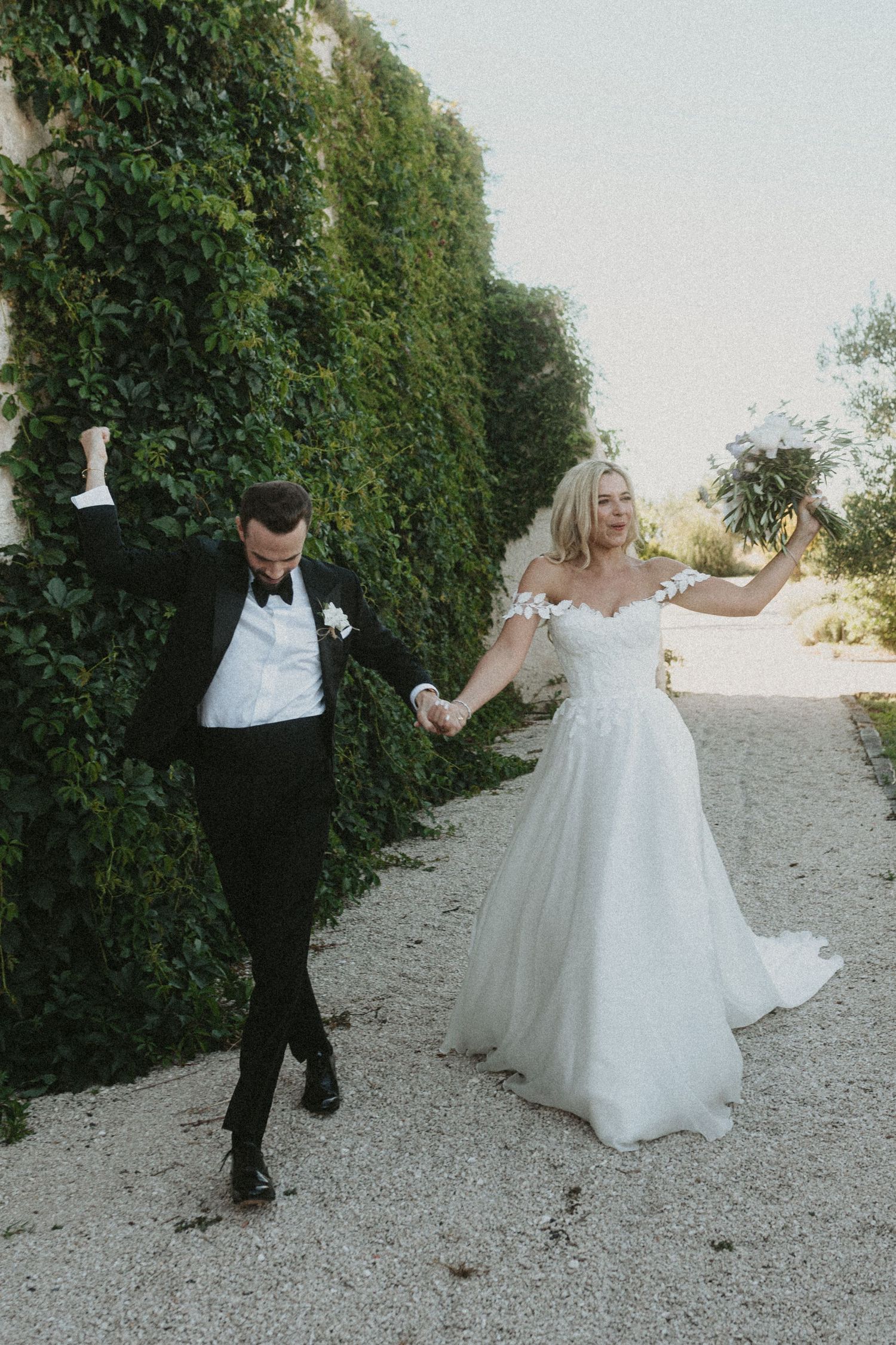 A couple in wedding attire walks hand in hand along a gravel path bordered by tall green hedges.