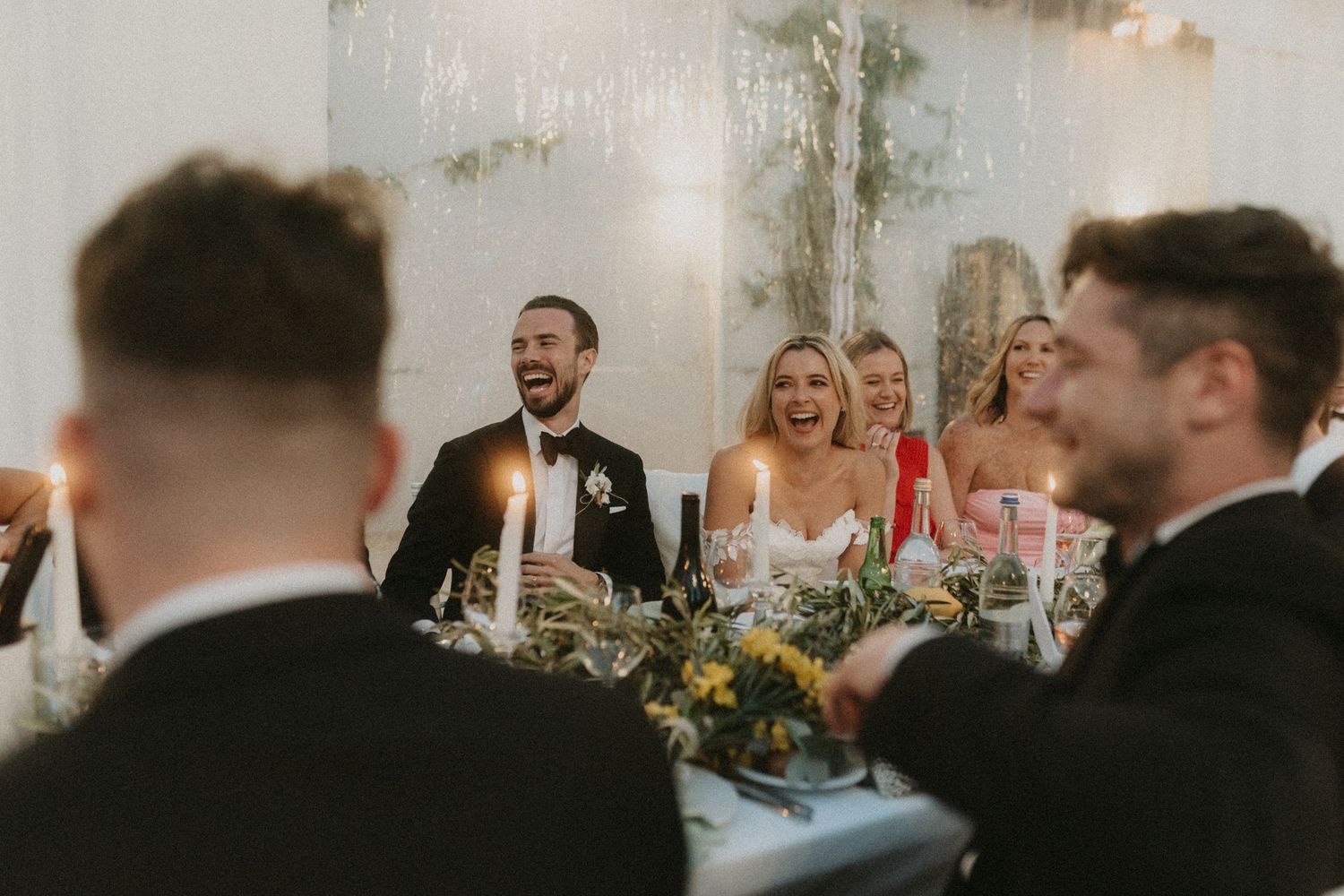 Wedding guests share laughter during reception dinner with elegant floral centerpieces and candlelight decor.