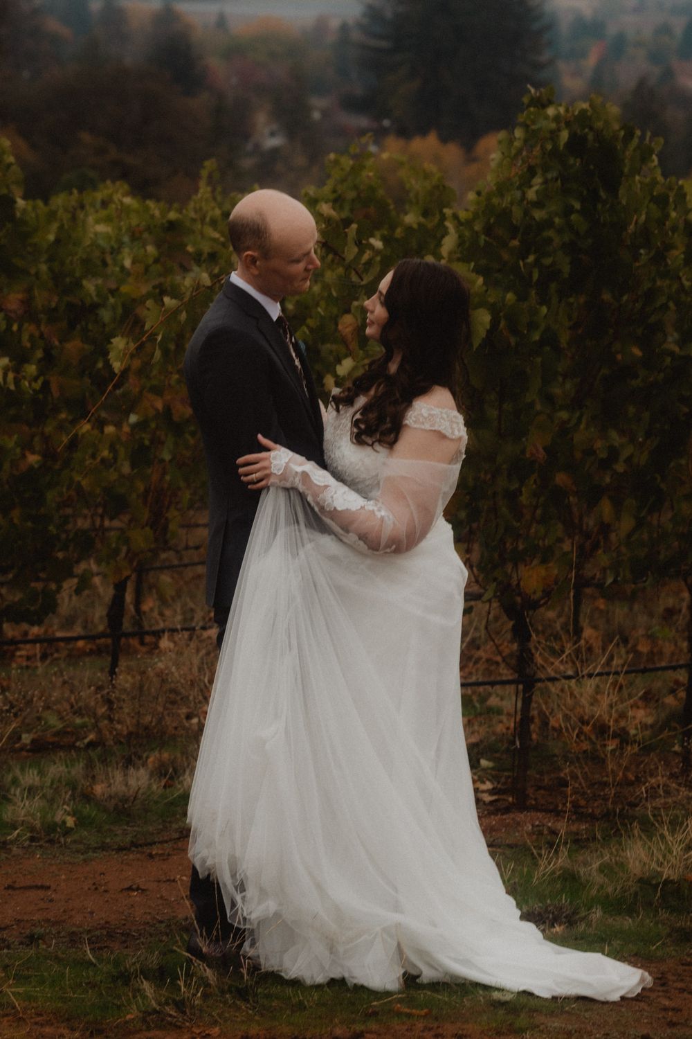 A couple in formal attire embraces in a scenic autumn vineyard setting at dusk.