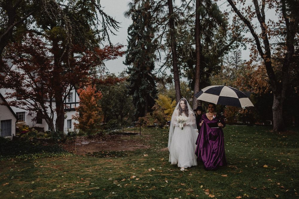 Wedding couple stands beneath autumn trees on a cloudy day with umbrellas and formal attire.