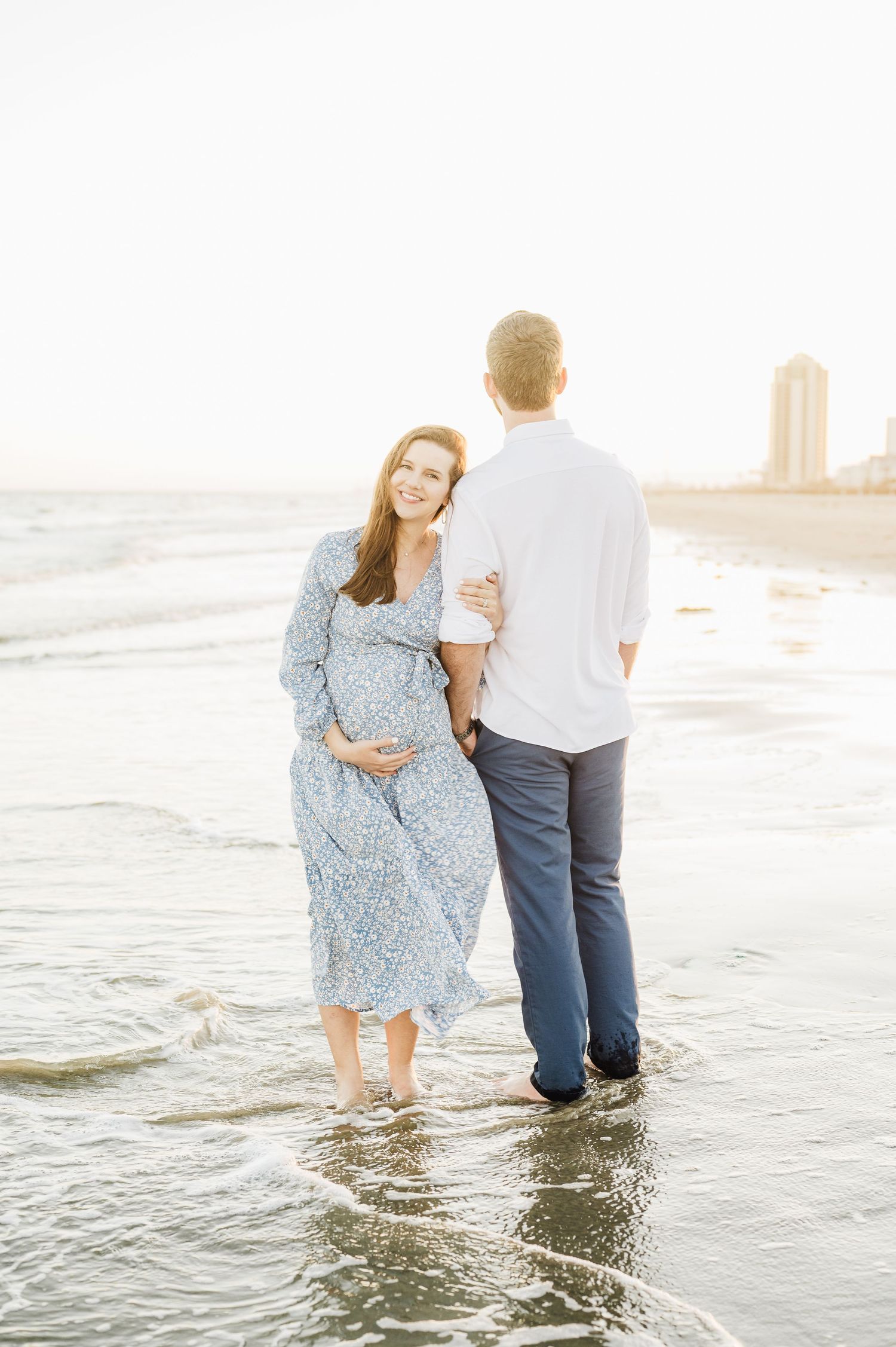 A couple stands in shallow ocean waves at sunset, with the woman wearing a flowing blue polka dot dress.