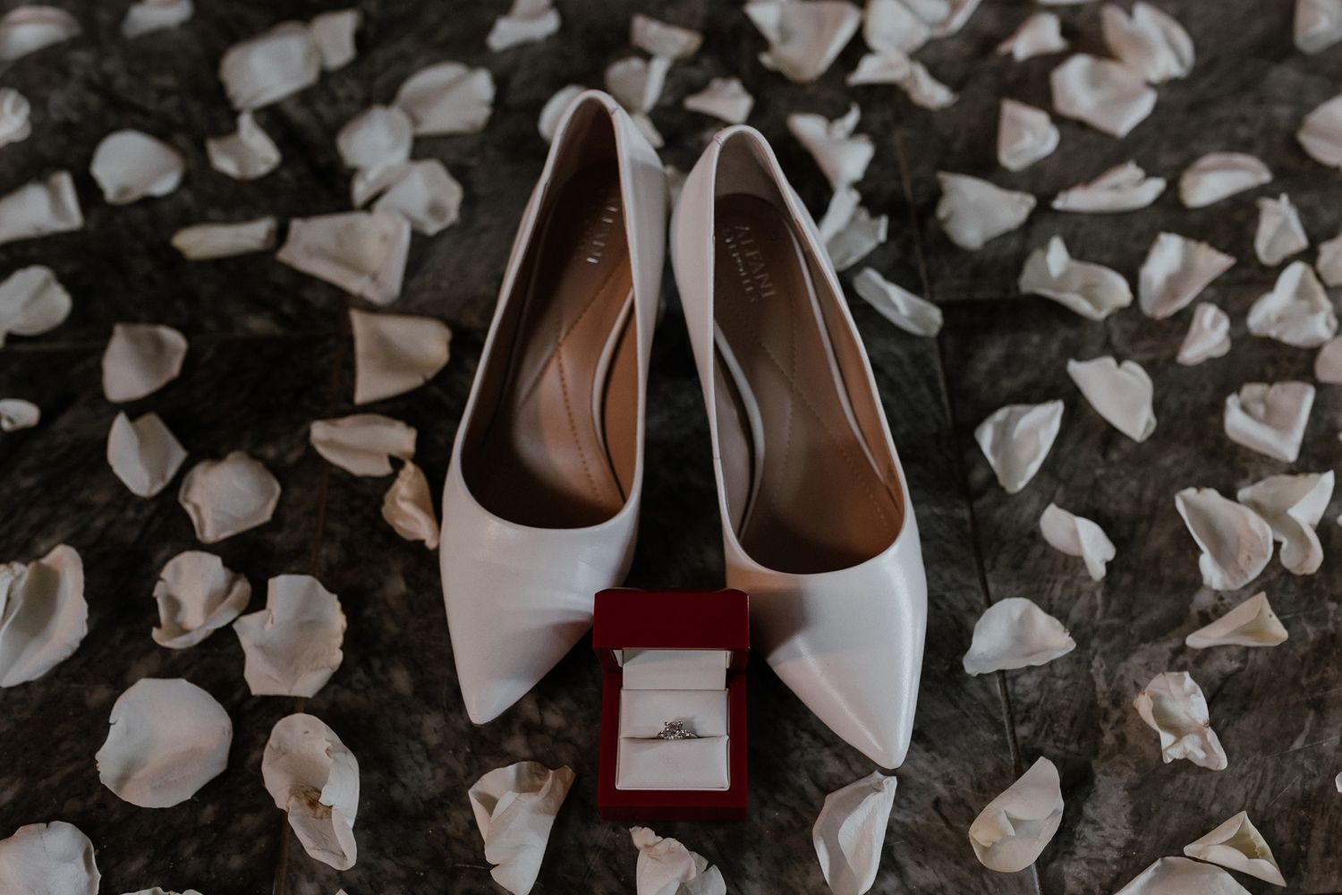 White bridal heels with engagement ring box on scattered rose petals against dark background.