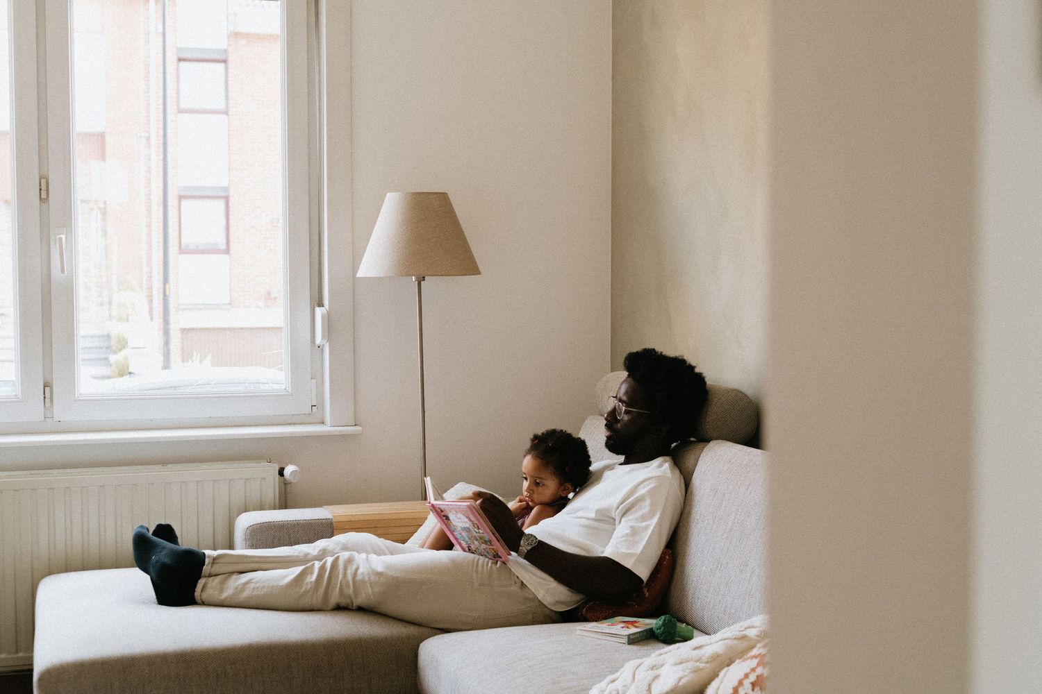 Person resting on a white couch in a bright, minimalist living room with natural light streaming through windows.