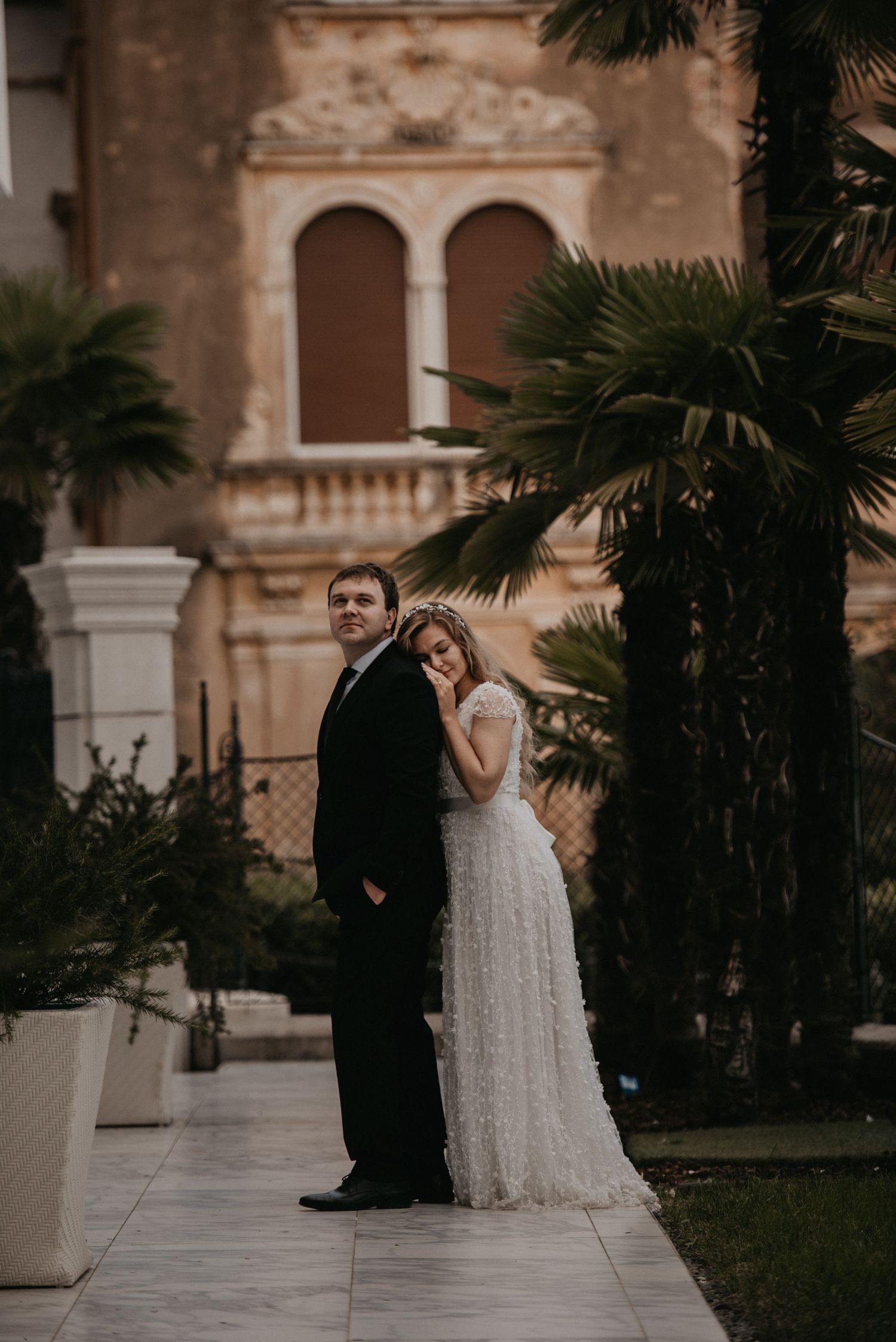 Elegant wedding couple poses in front of Mediterranean architecture with palm trees.