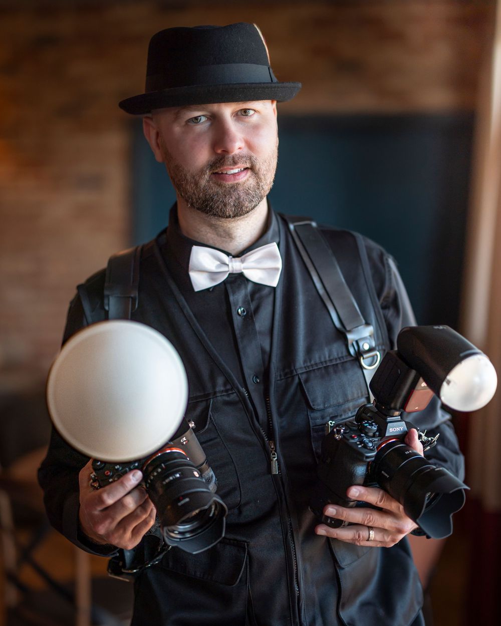 Professional photographer in bow tie and hat holding camera equipment and lighting gear.
