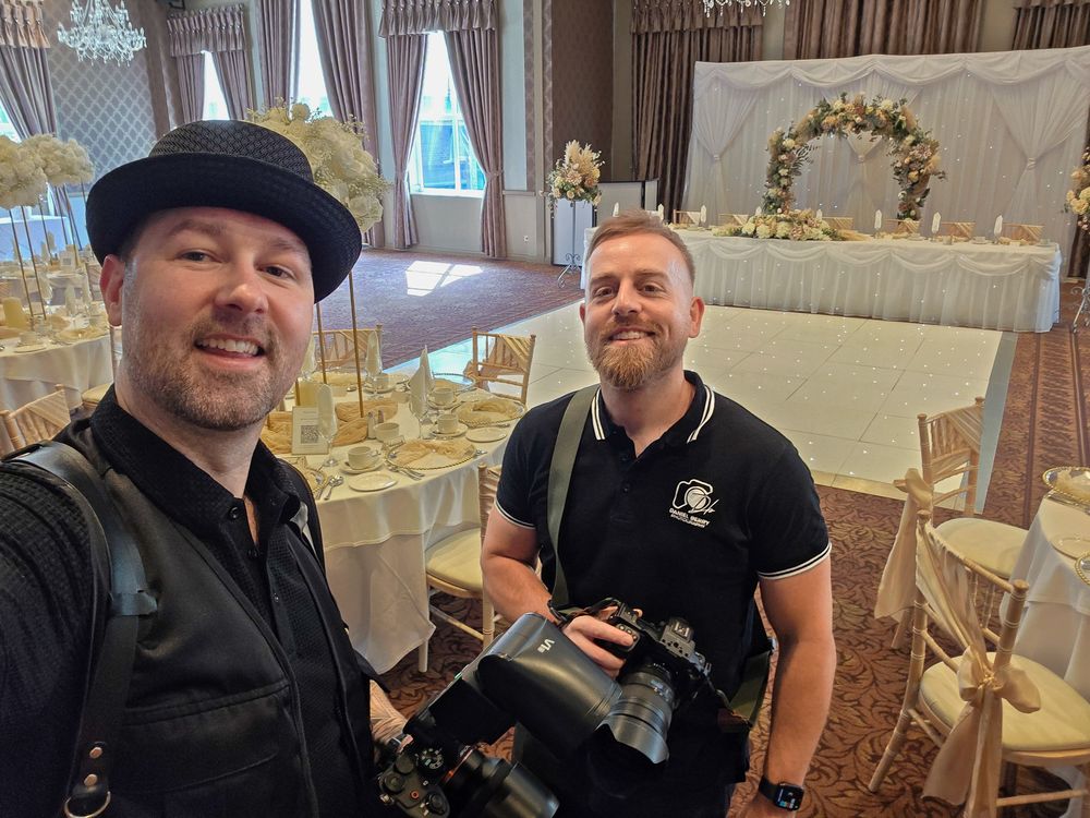 Two photographers smile for a selfie inside an elegantly decorated wedding venue with floral arrangements.