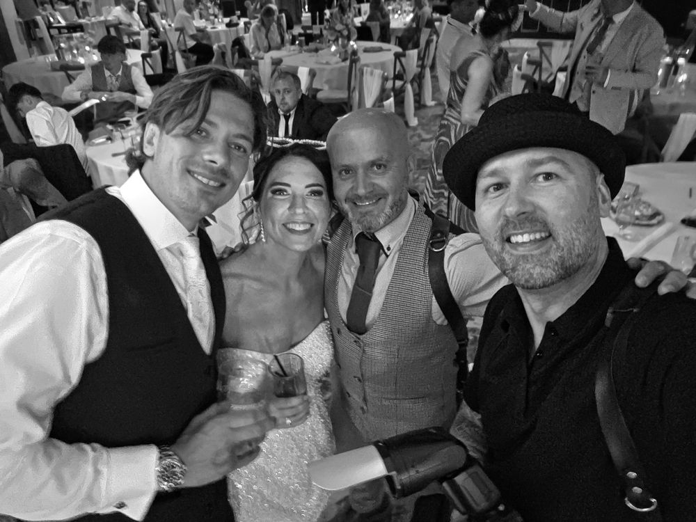 Black and white wedding photo of bride and groom posing with two guests at a festive reception dinner.